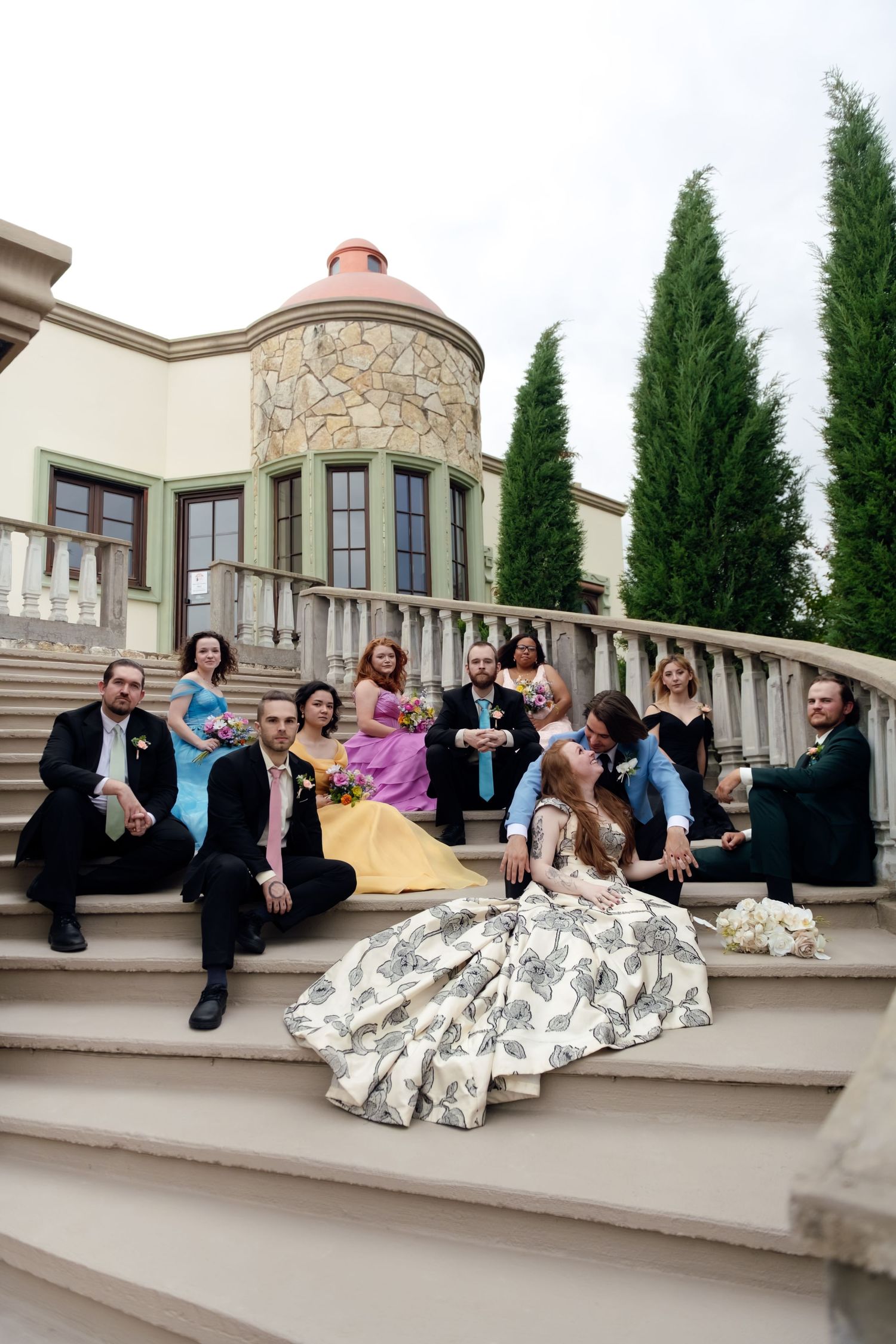 Wedding party posed on grand stone steps with cypress trees in background.