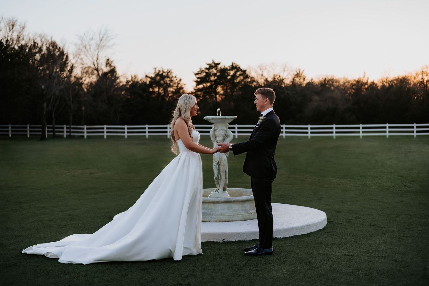 A wedding ceremony takes place at sunset on a grassy field with white fencing in the background.