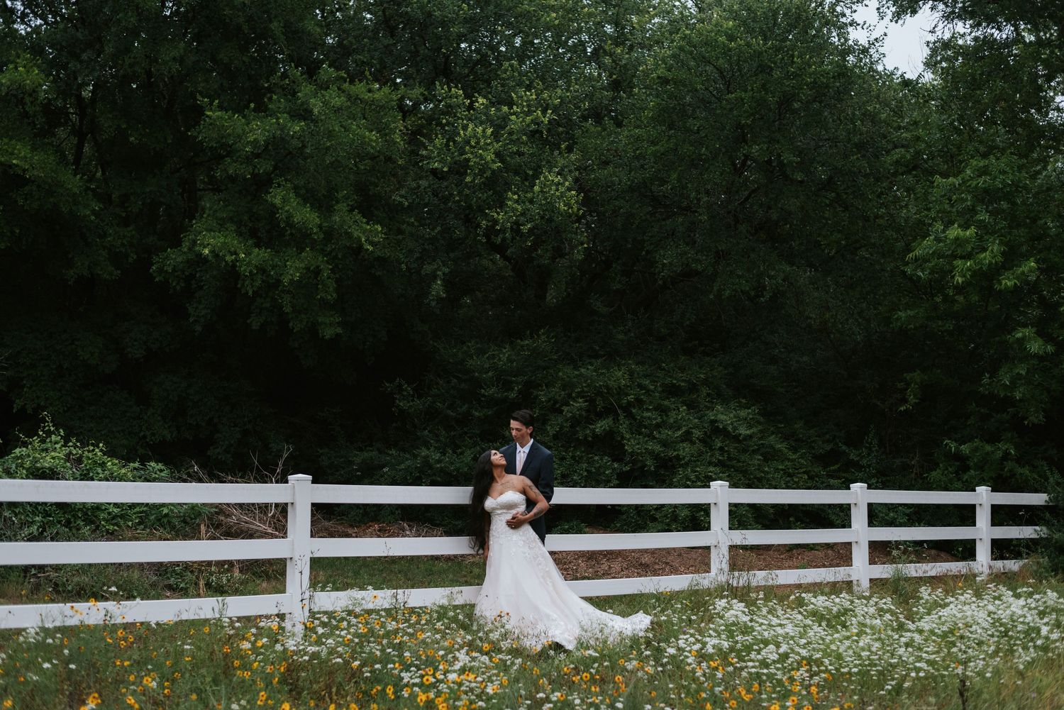 Wedding couple posing by white fence with wildflowers in countryside setting.