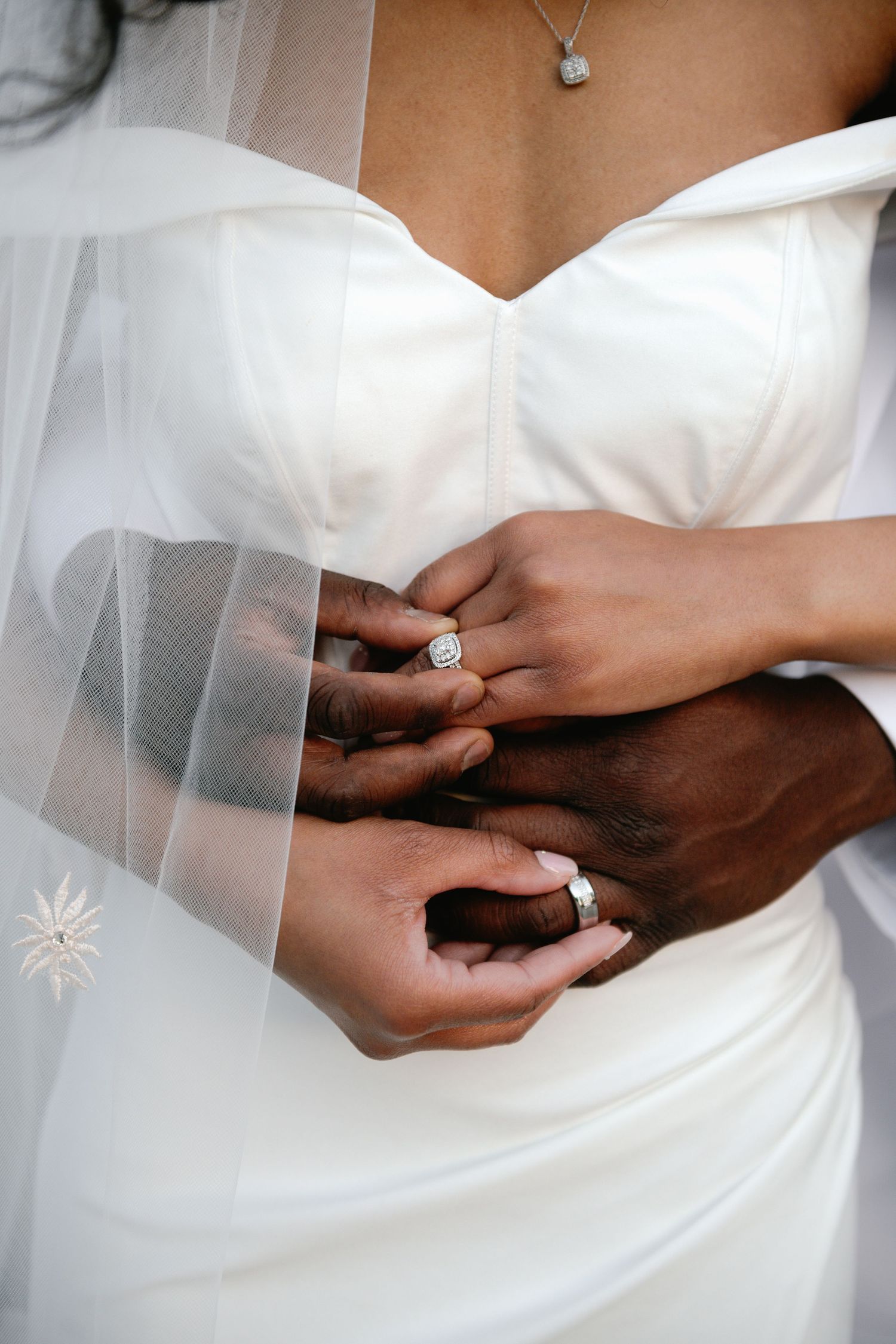Close-up of hands with wedding rings against white wedding dress and veil.