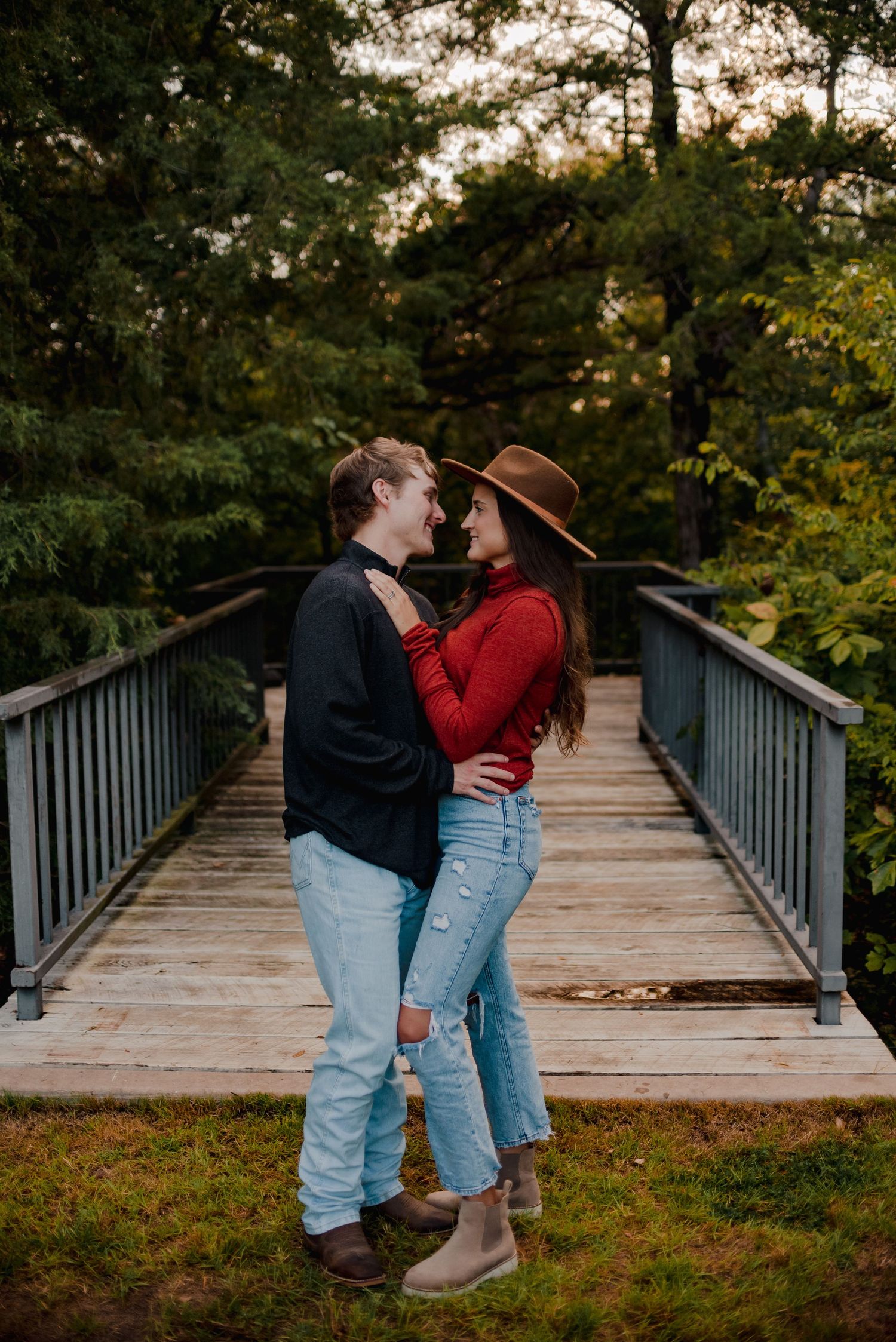 A couple in casual attire embraces on a wooden footbridge in a park setting.