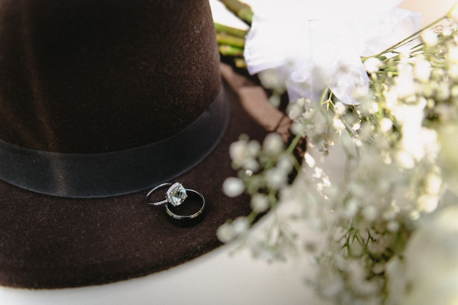 Brown fedora hat with wedding rings and white wildflowers, elegant bridal accessories flat lay photography.