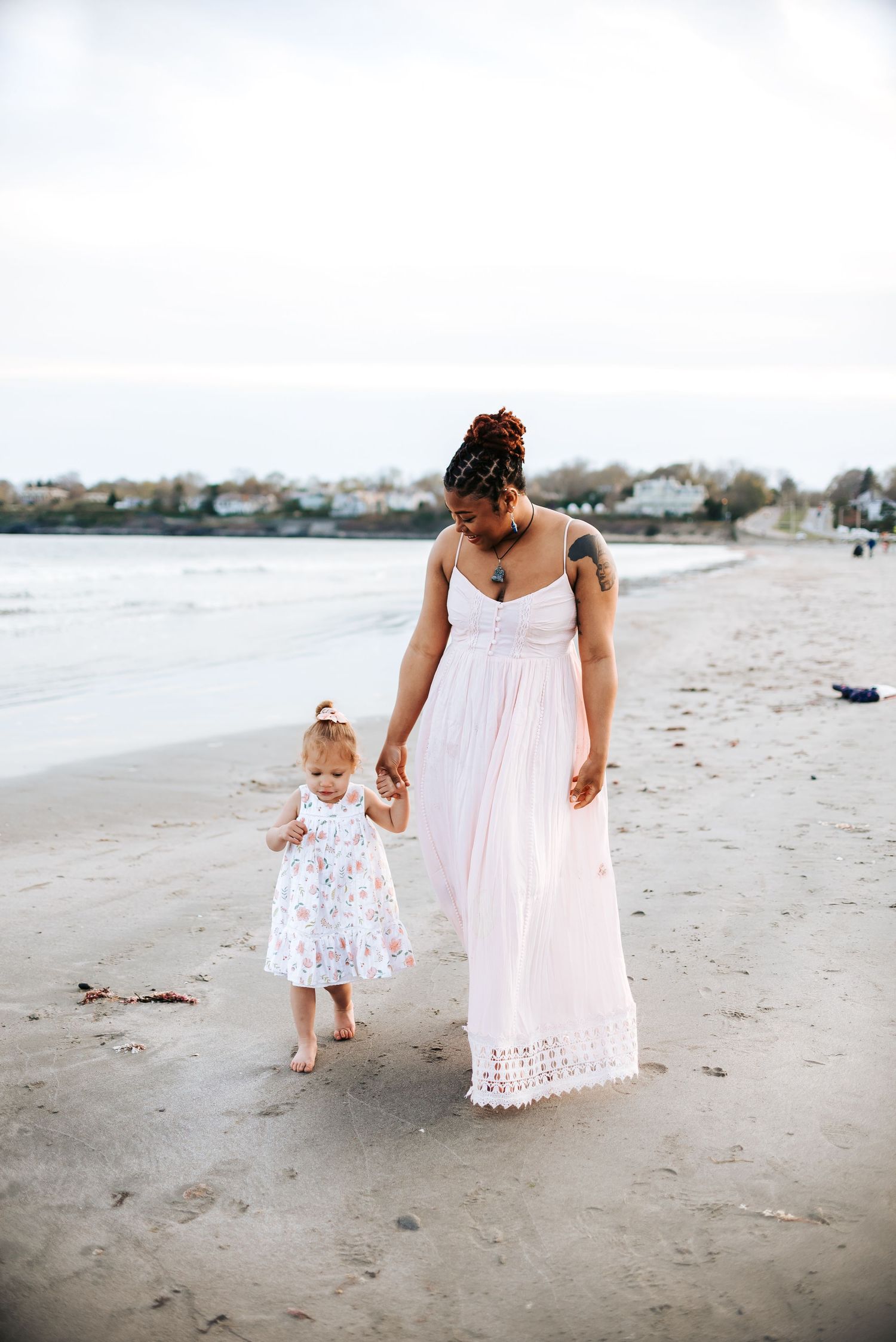 Mother and daughter walking together on sandy beach in matching white dresses.
