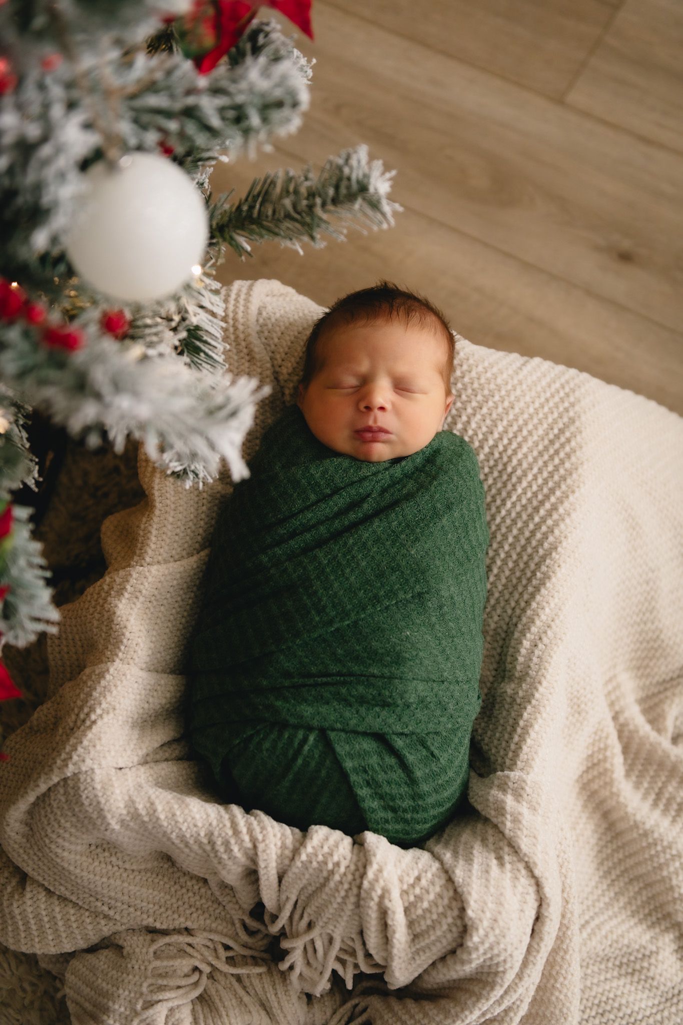 A newborn baby wrapped in green swaddle sleeps peacefully next to a Christmas tree.