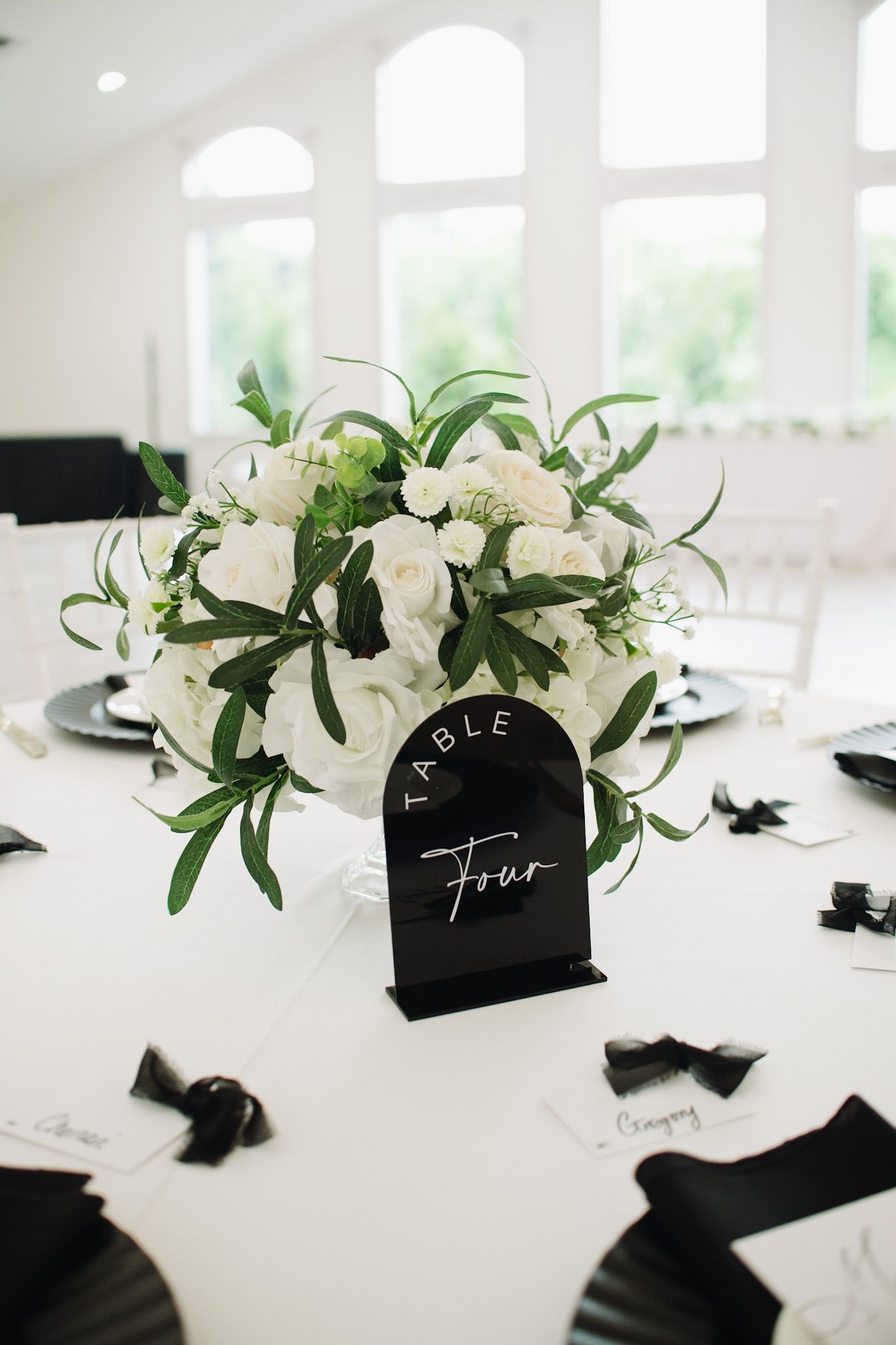Modern wedding table setting with white roses and black place cards in bright indoor venue.