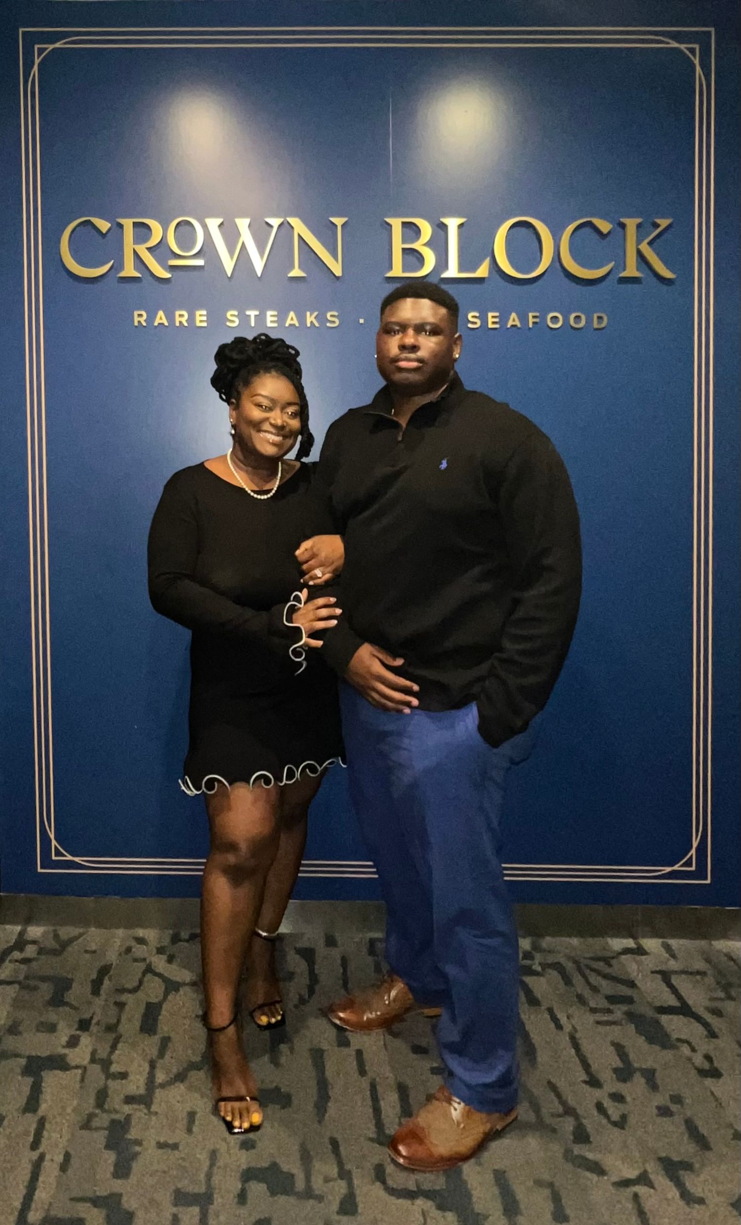 Couple taking photos at Crown Block restaurant with blue wall and gold logo backdrop.