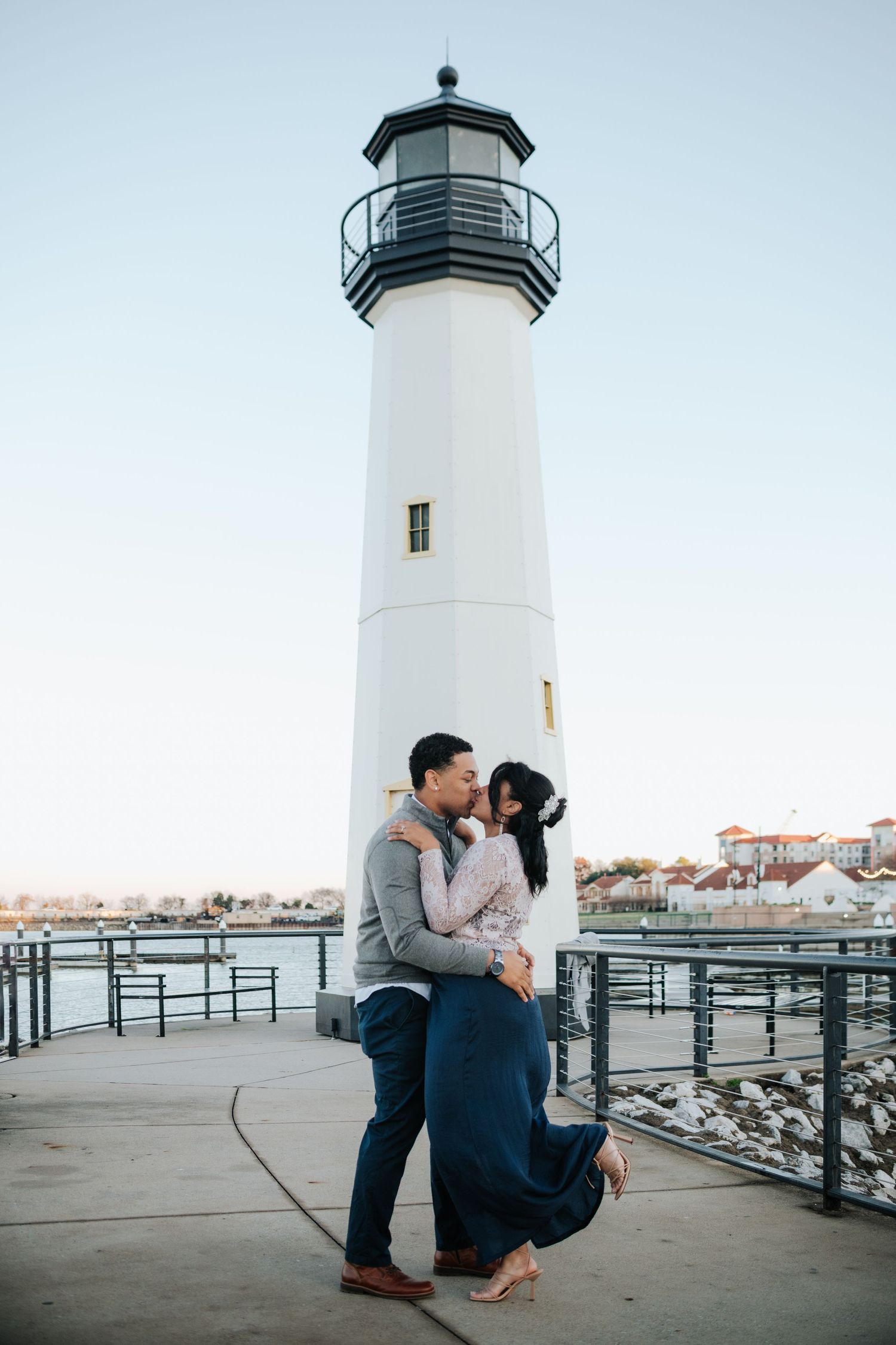 Couple shares intimate moment near white lighthouse at sunset.