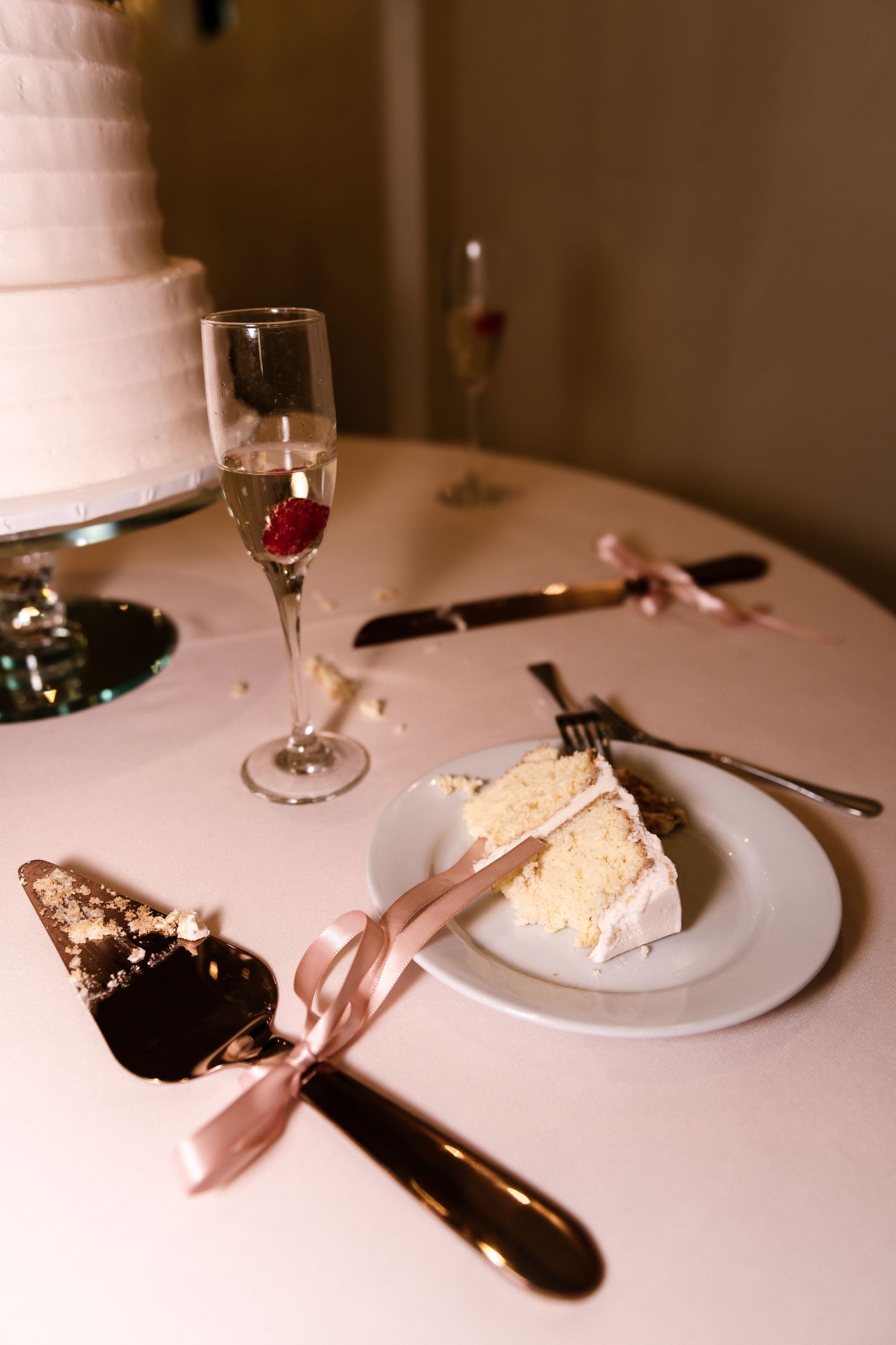 Wedding cake slice and champagne glass on white tablecloth with rose gold utensils.