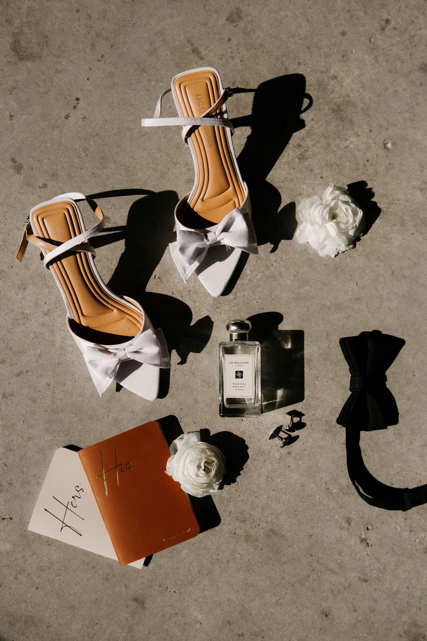 Wedding accessories including white heels, perfume bottle, black bow tie and white flowers arranged on concrete floor.