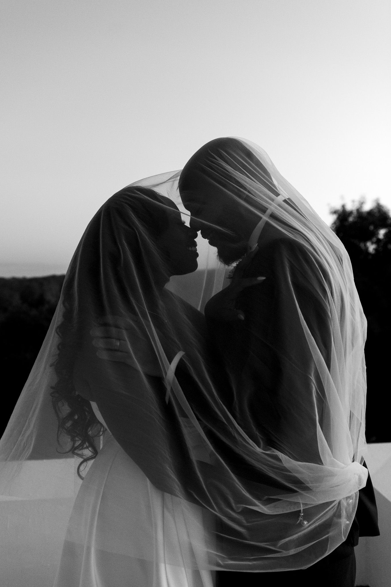 Romantic black and white portrait of couple sharing intimate moment under wedding veil at sunset.