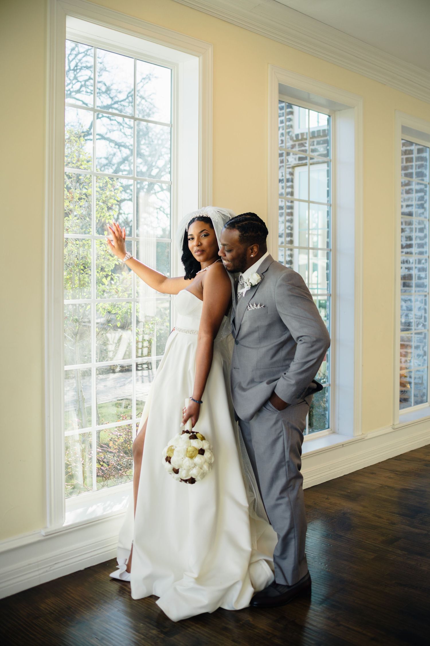 A couple takes a selfie by large windows in formal wedding attire.