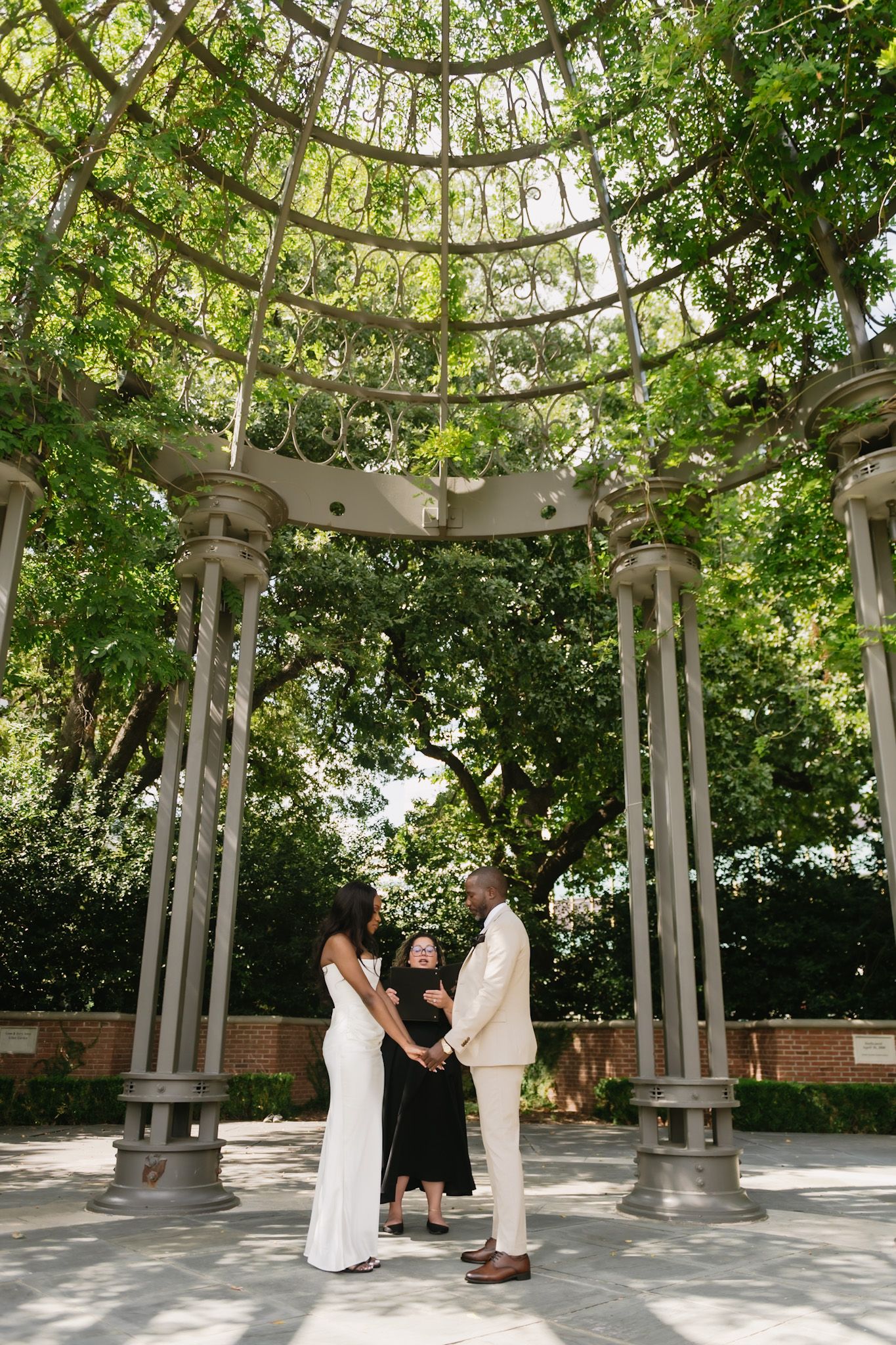 Wedding ceremony under white pergola surrounded by lush green trees.