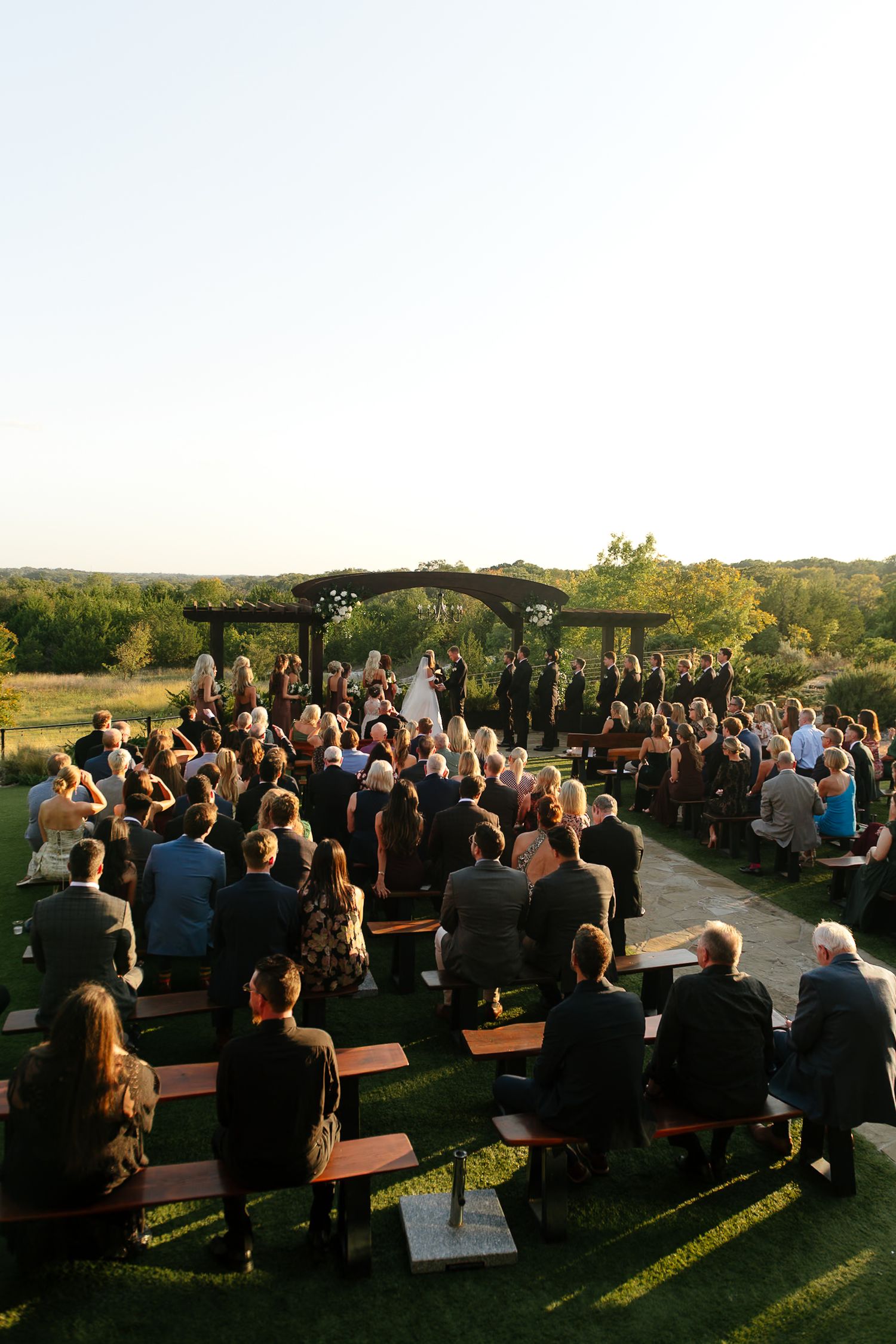 Outdoor wedding ceremony setup with wooden benches and guests seated in a natural landscape.