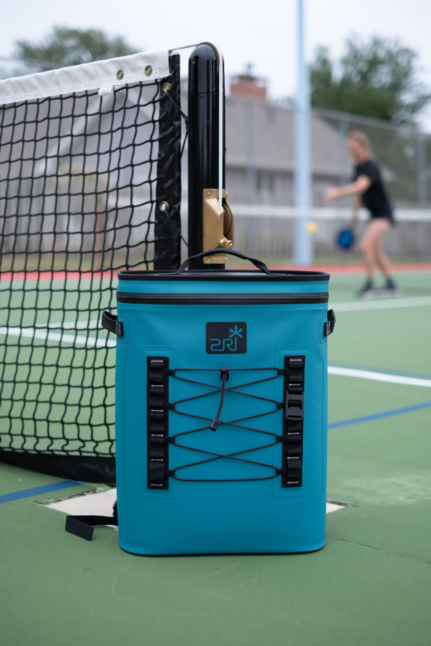 A blue PRI cooler is featured in front of a woman playing pickleball.