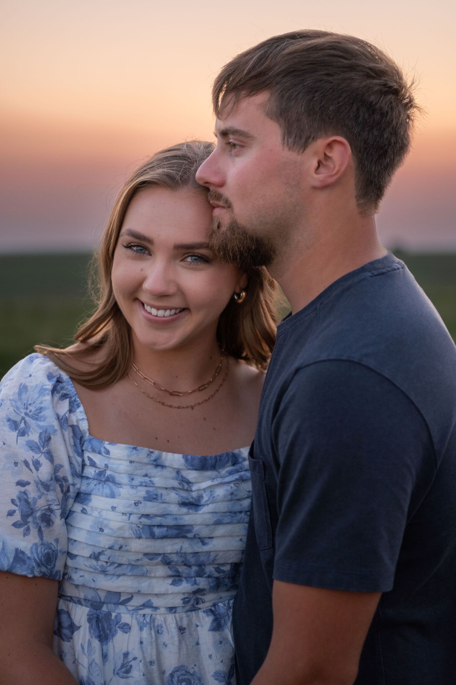 A woman looking directly at the camera, a man looking off into the distance while the sun sets behind them during a couples session.