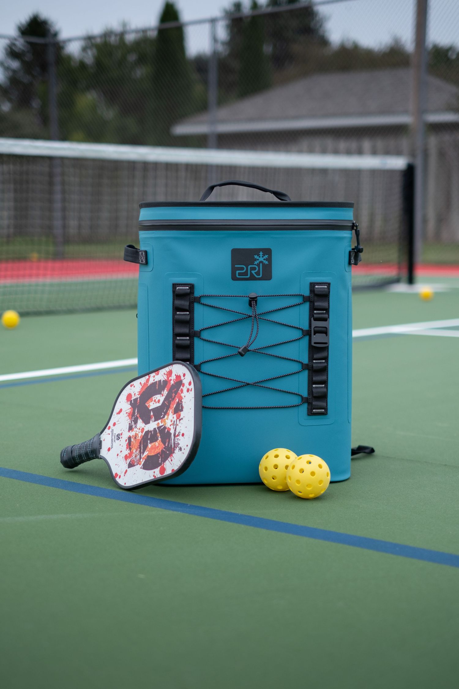 A blue PRI cooler set on a pickleball court with a paddle and pickleballs.