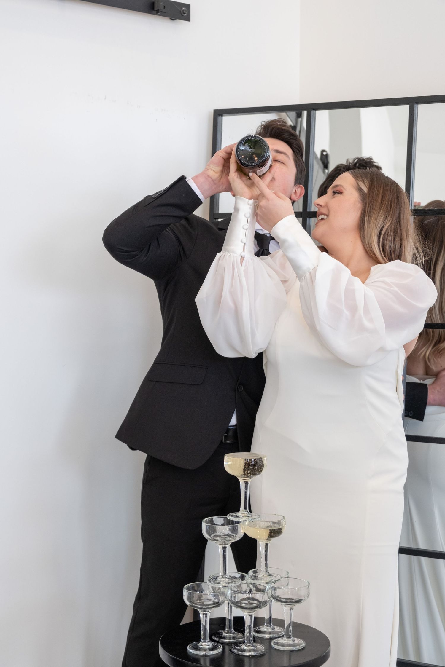 A bride and groom share a celebratory drink behind a champagne tower.