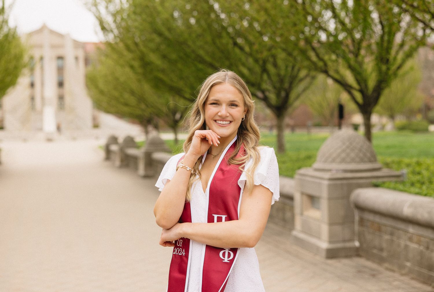 Graduate wearing white dress and red sorority stole poses on spring campus path.