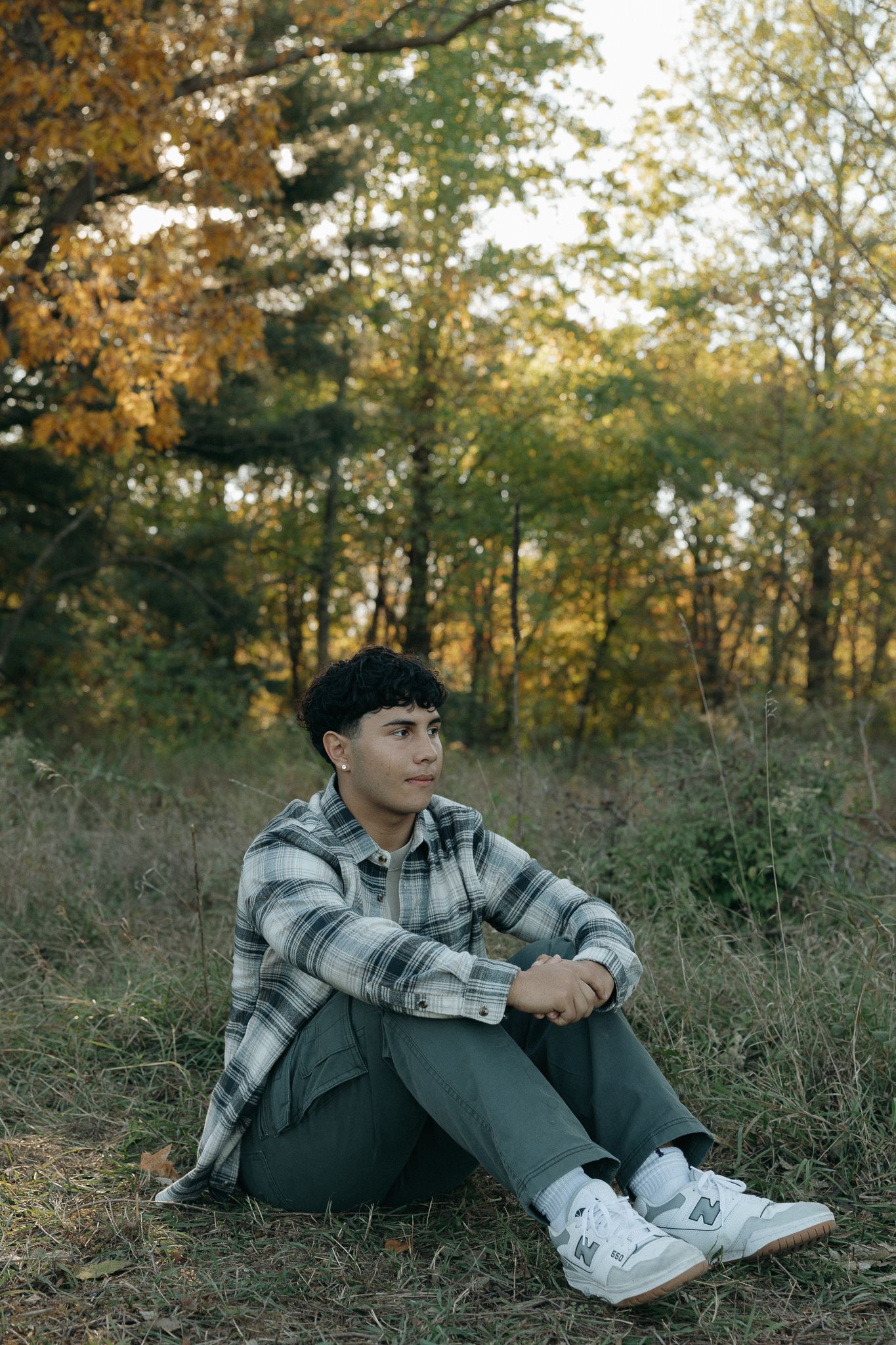 Person in casual attire sits on ground surrounded by fall foliage.