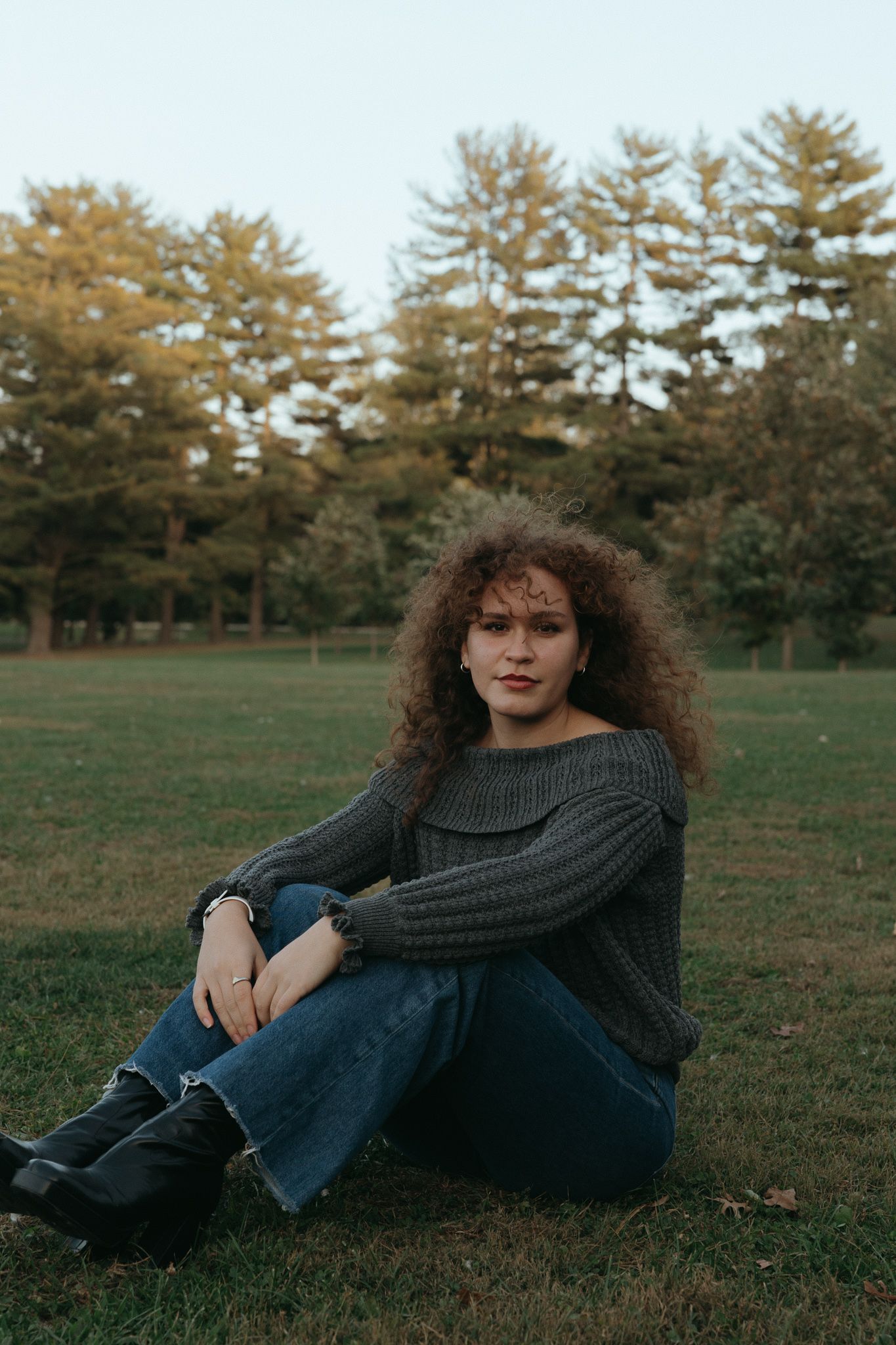 A person in a gray sweater and jeans sits on grass with autumn trees in the background.