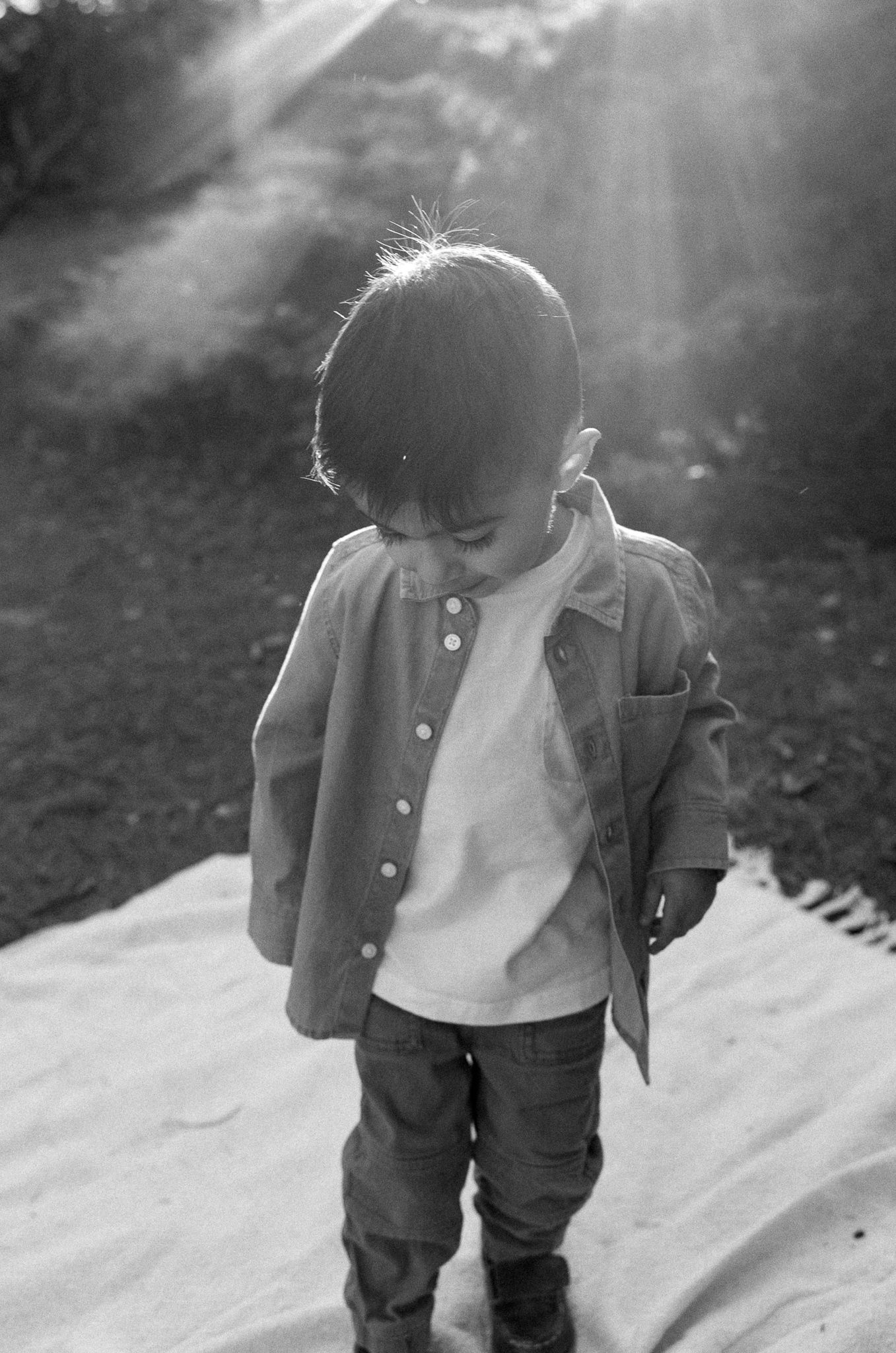 A child in a button-up jacket looks down while standing in dramatic backlit sunlight.