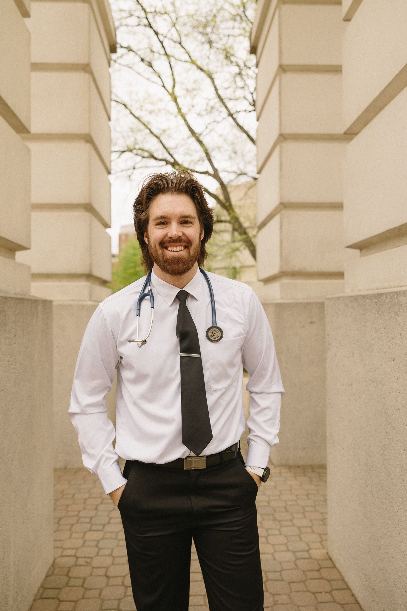 A medical professional in white shirt and tie stands between stone columns on campus.