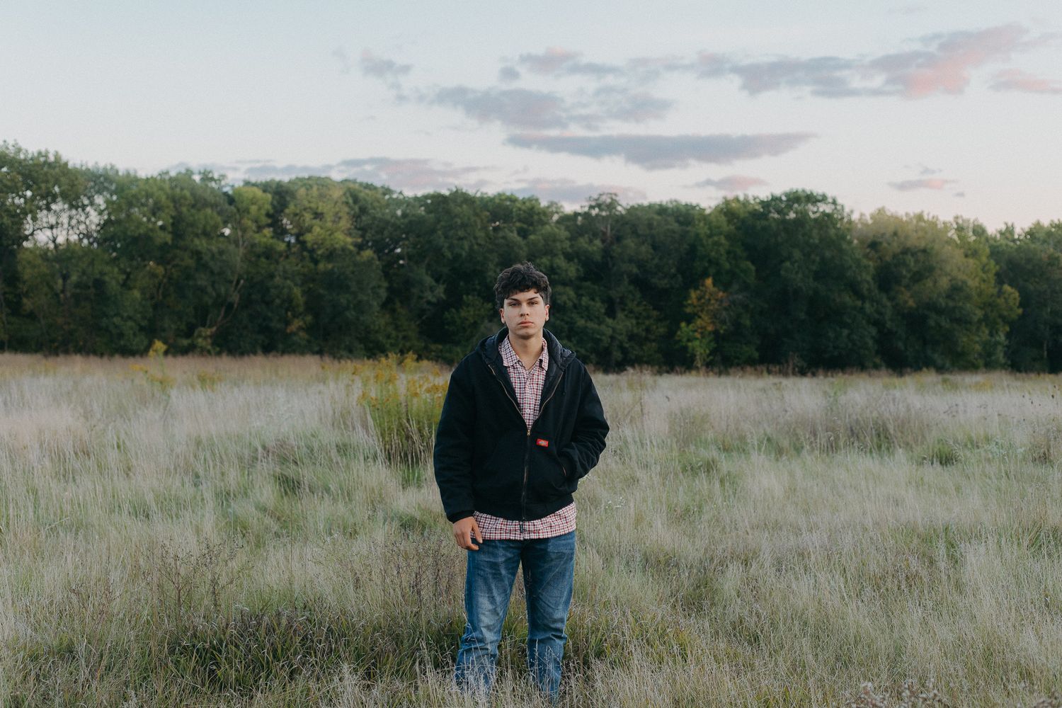 Person in dark jacket standing in tall grass field during golden hour.