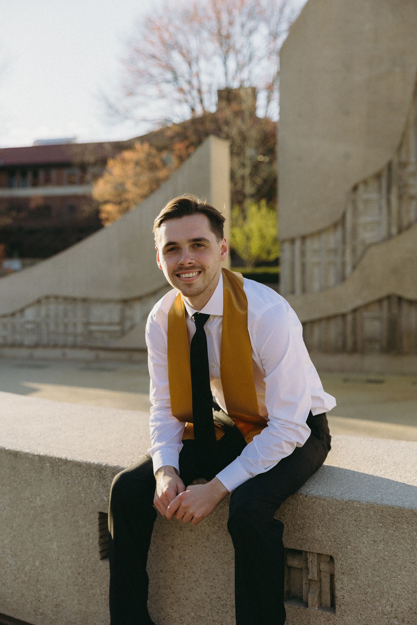 A graduate in business attire with gold stole sits on a concrete wall on campus.