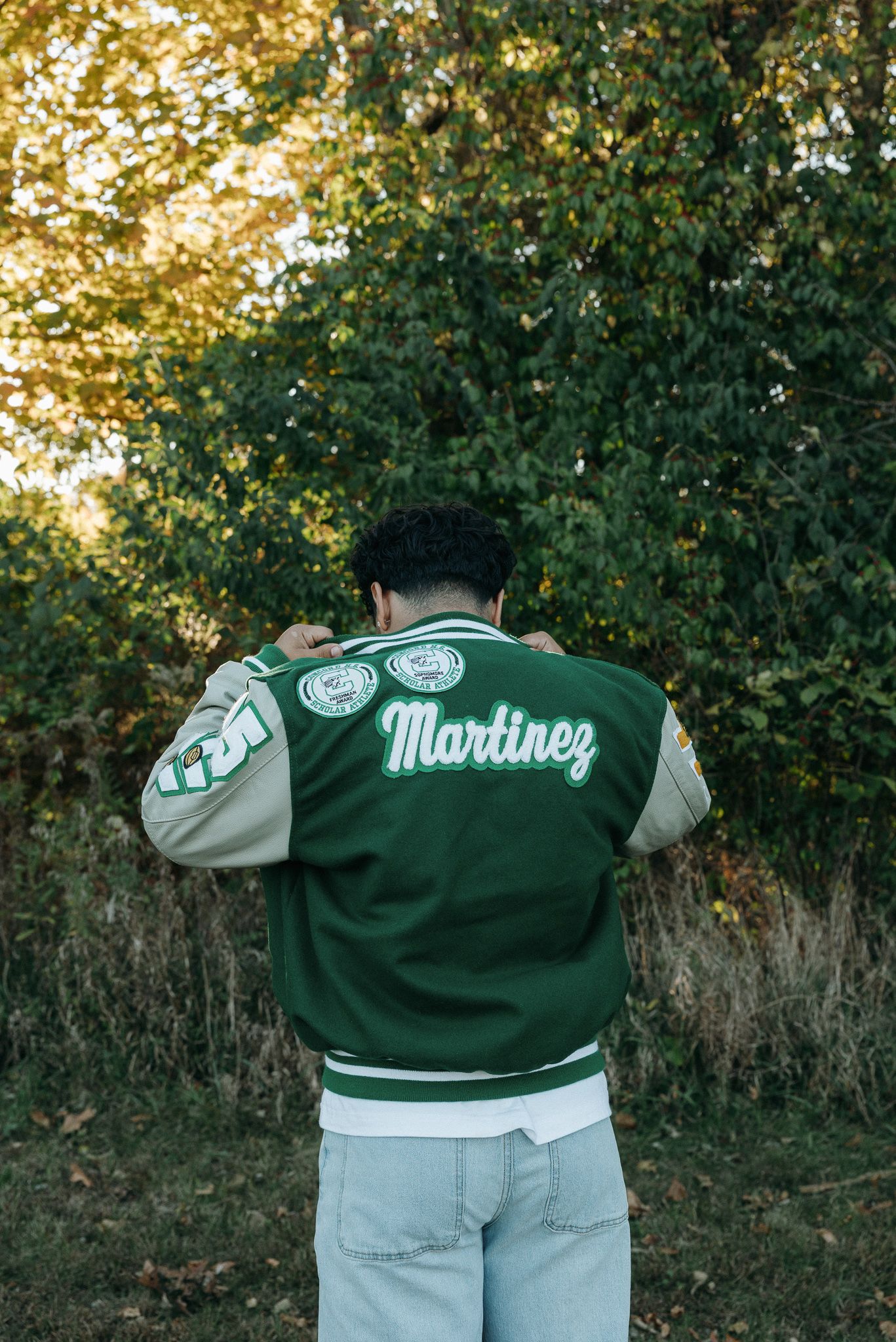 Rear view of varsity jacket with 'Martinez' lettering against autumn foliage background.