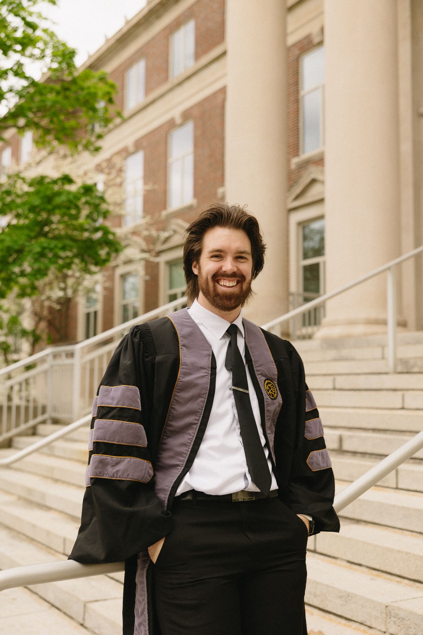 A doctoral graduate in academic regalia poses on the steps of a Purdue University building.