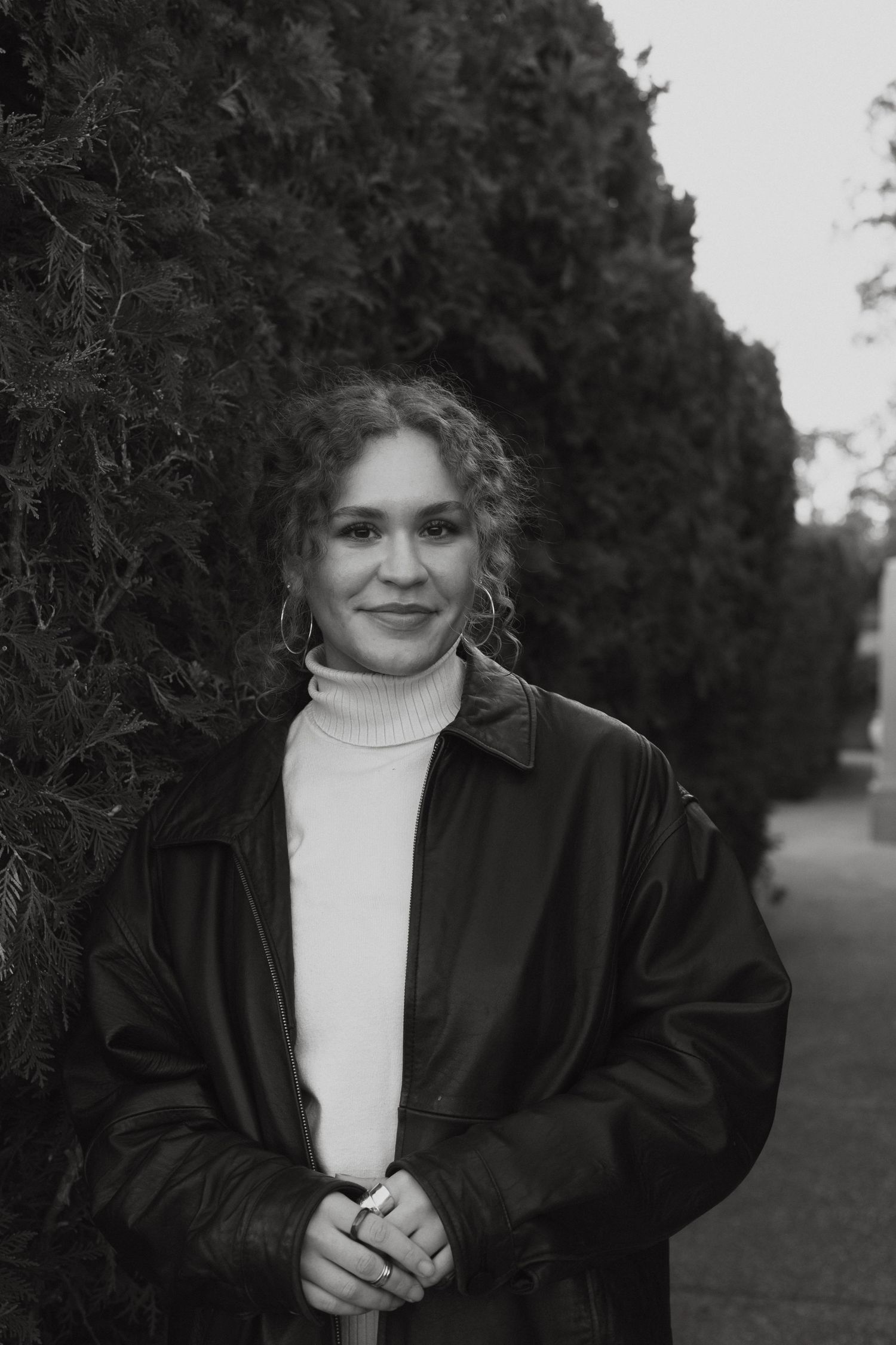 Portrait of someone in turtleneck and jacket against evergreen hedge.