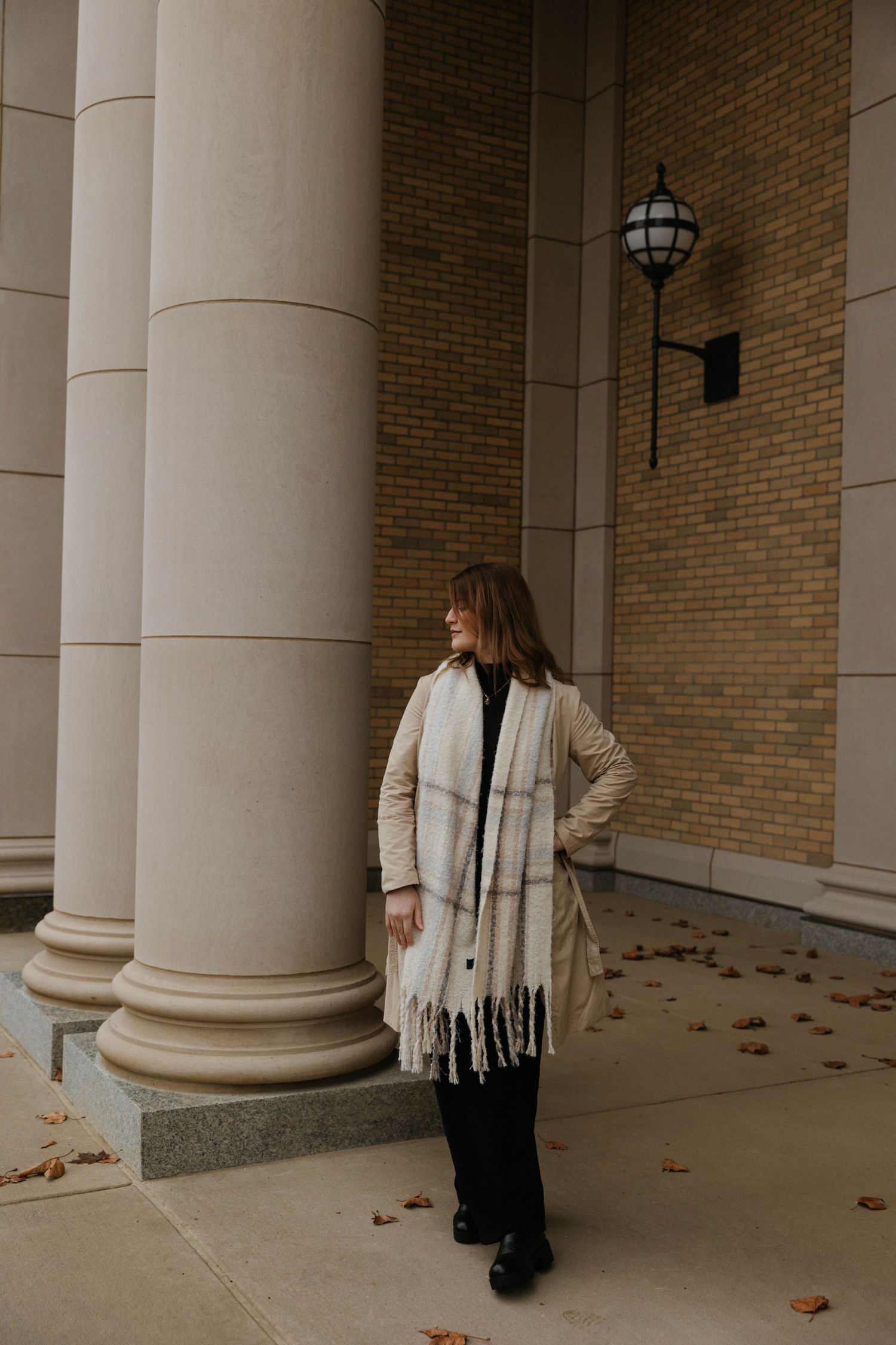 Person in cream colored coat and scarf standing between architectural columns.