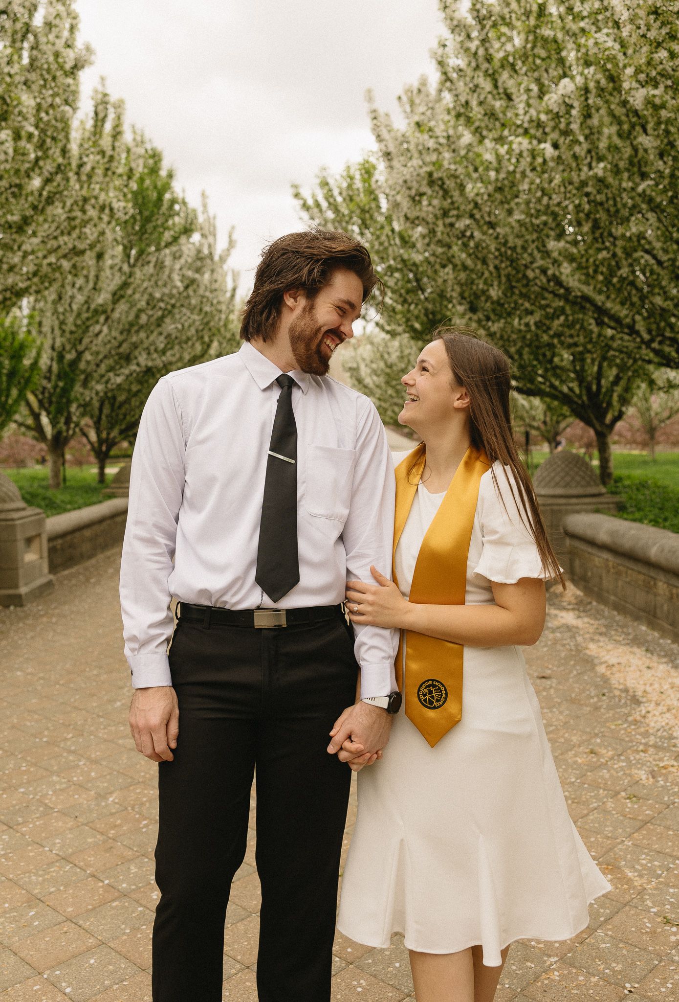 A couple in graduation attire share a moment on a tree-lined path at Purdue University.