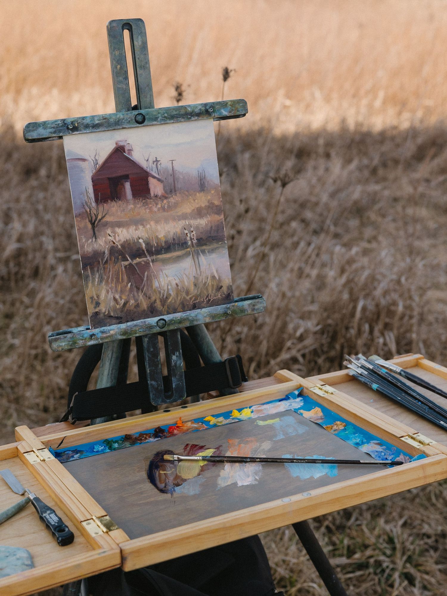 A painting of a red barn sits on an easel with painting supplies arranged on a wooden palette.