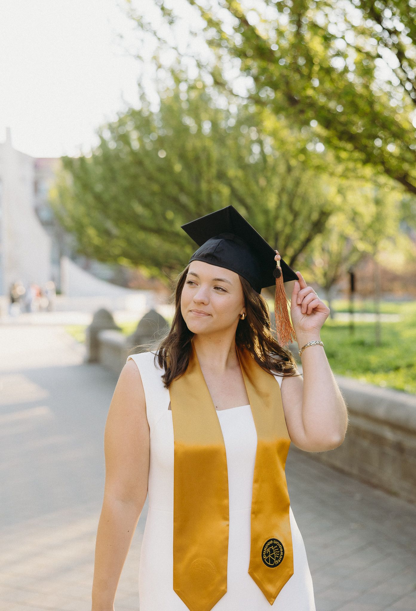 Graduate in white dress and gold stole poses outside on sunny spring day with graduation cap.