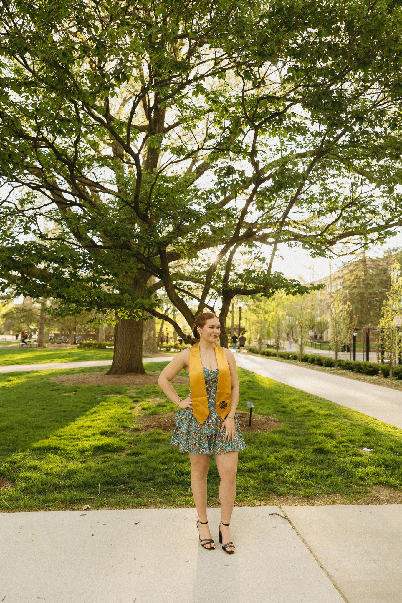 Graduate in yellow stole and patterned dress standing on campus lawn with large tree.
