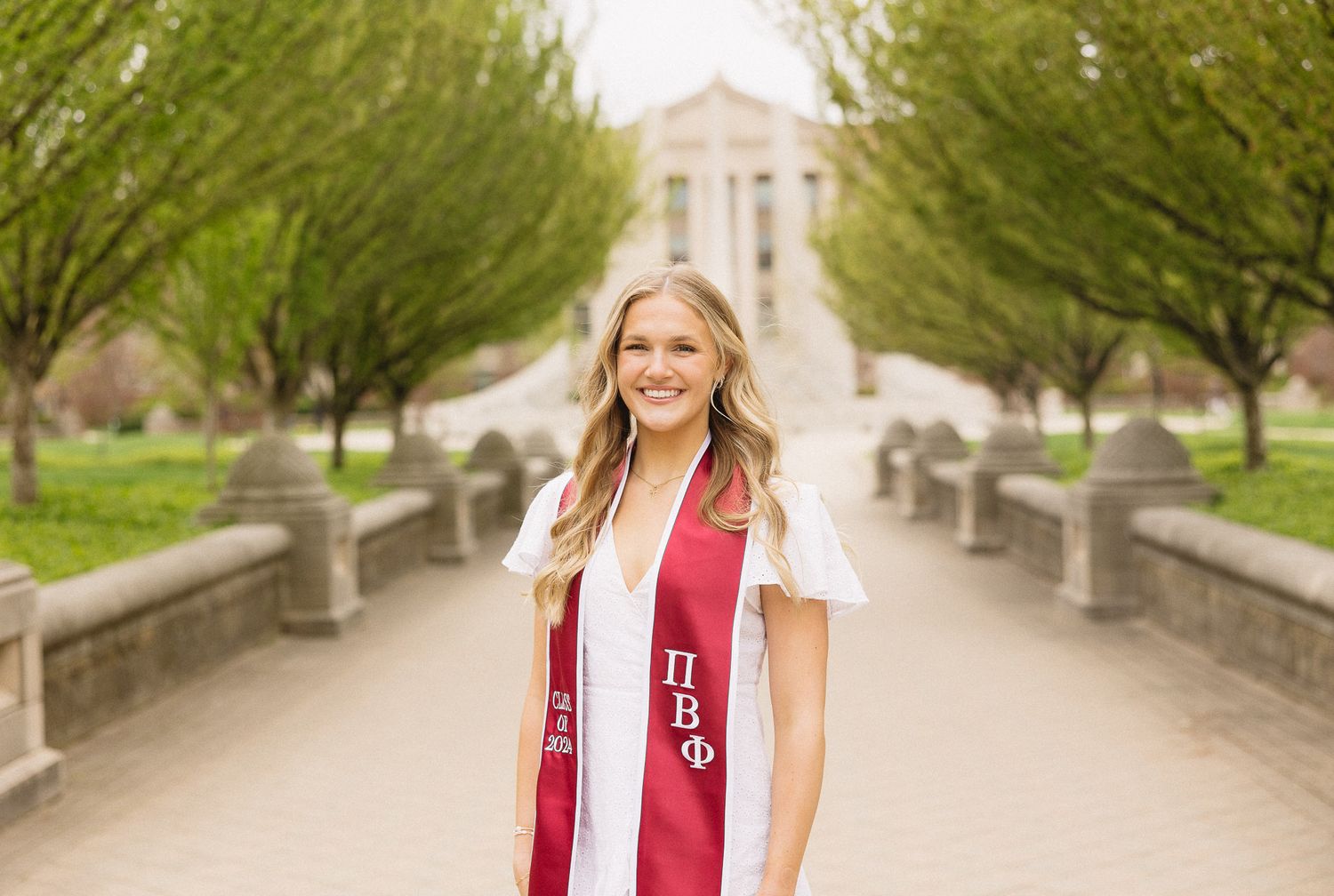 Graduate in white dress and red stole poses on tree-lined campus walkway.