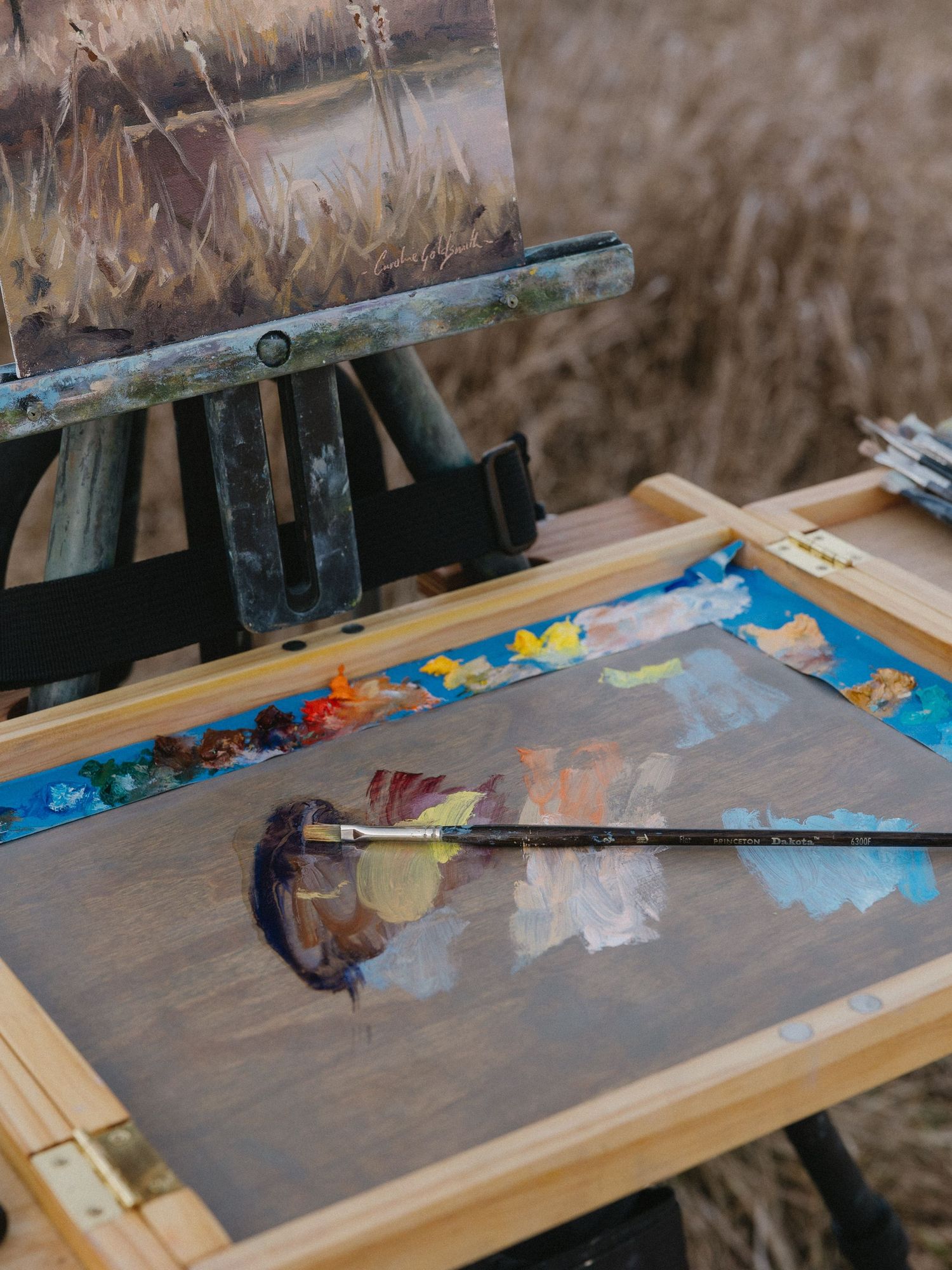 Close-up of an artist's palette with paint and brushes next to a canvas on an outdoor easel.