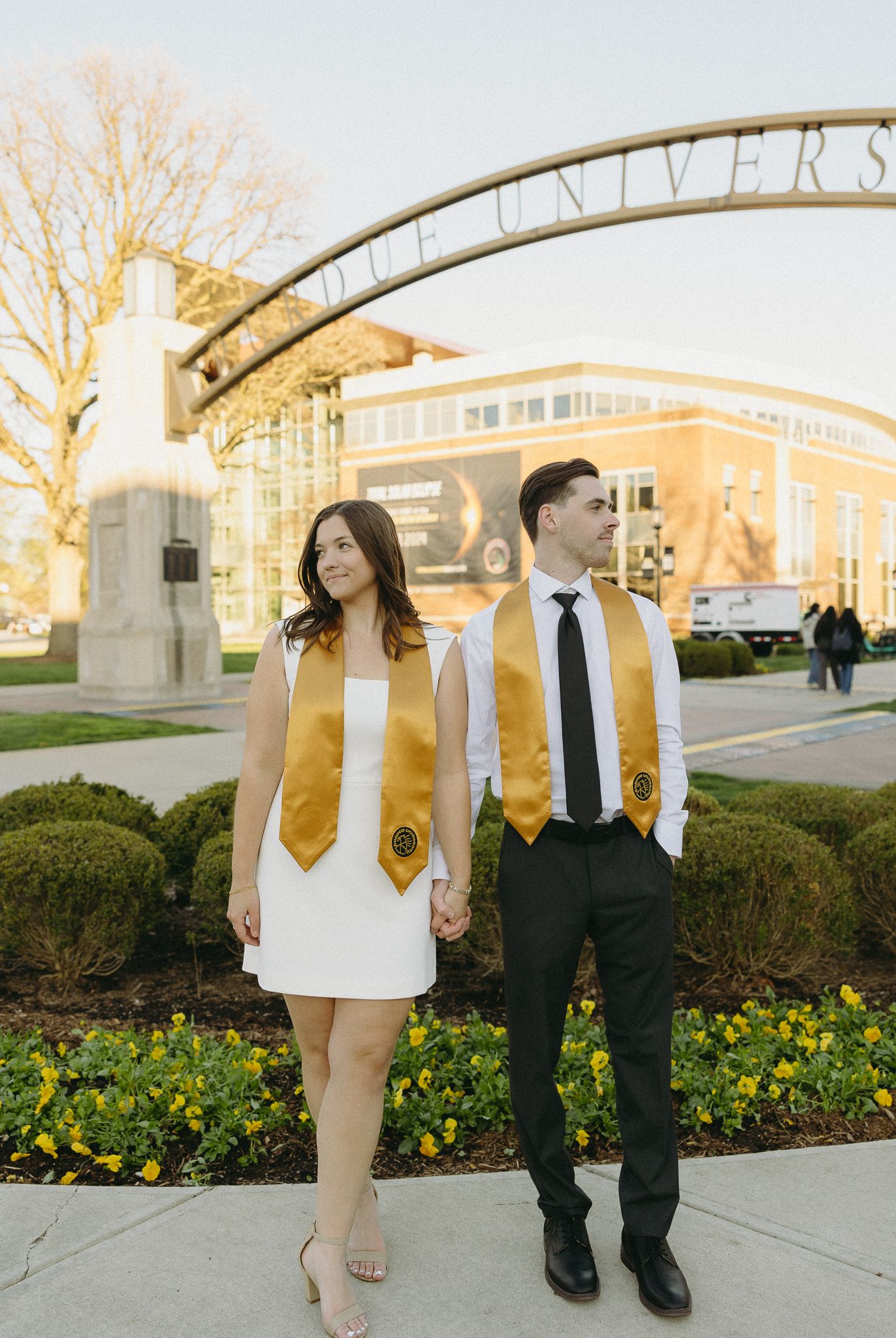 Graduates pose under university entrance arch wearing coordinating gold and white attire.