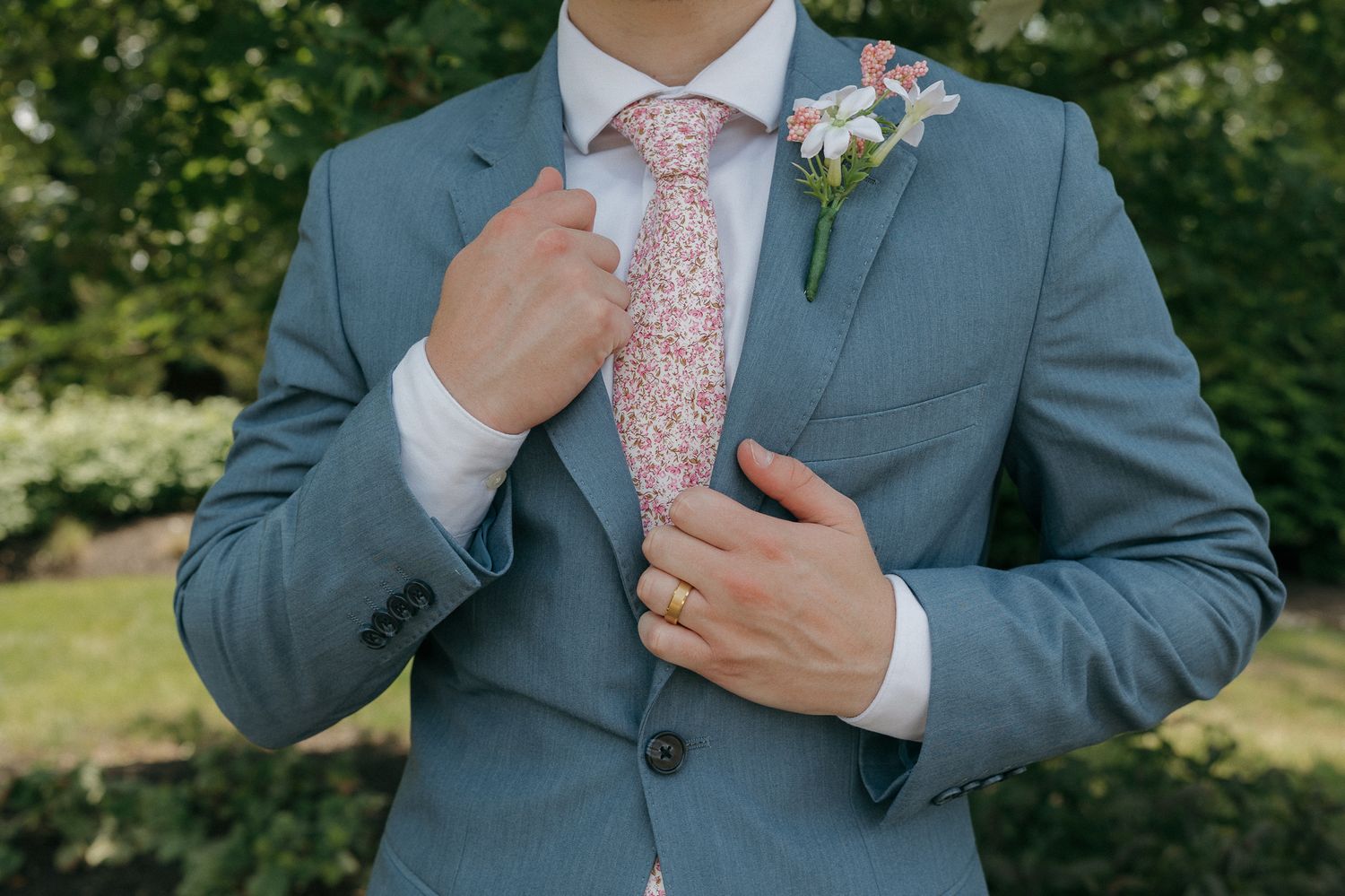Close-up of groom in gray suit adjusting pink floral tie with boutonniere.