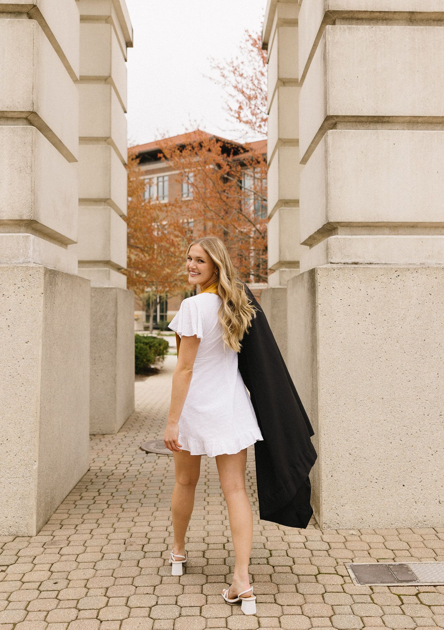 Graduate in white dress poses between stone pillars on textured brick walkway.