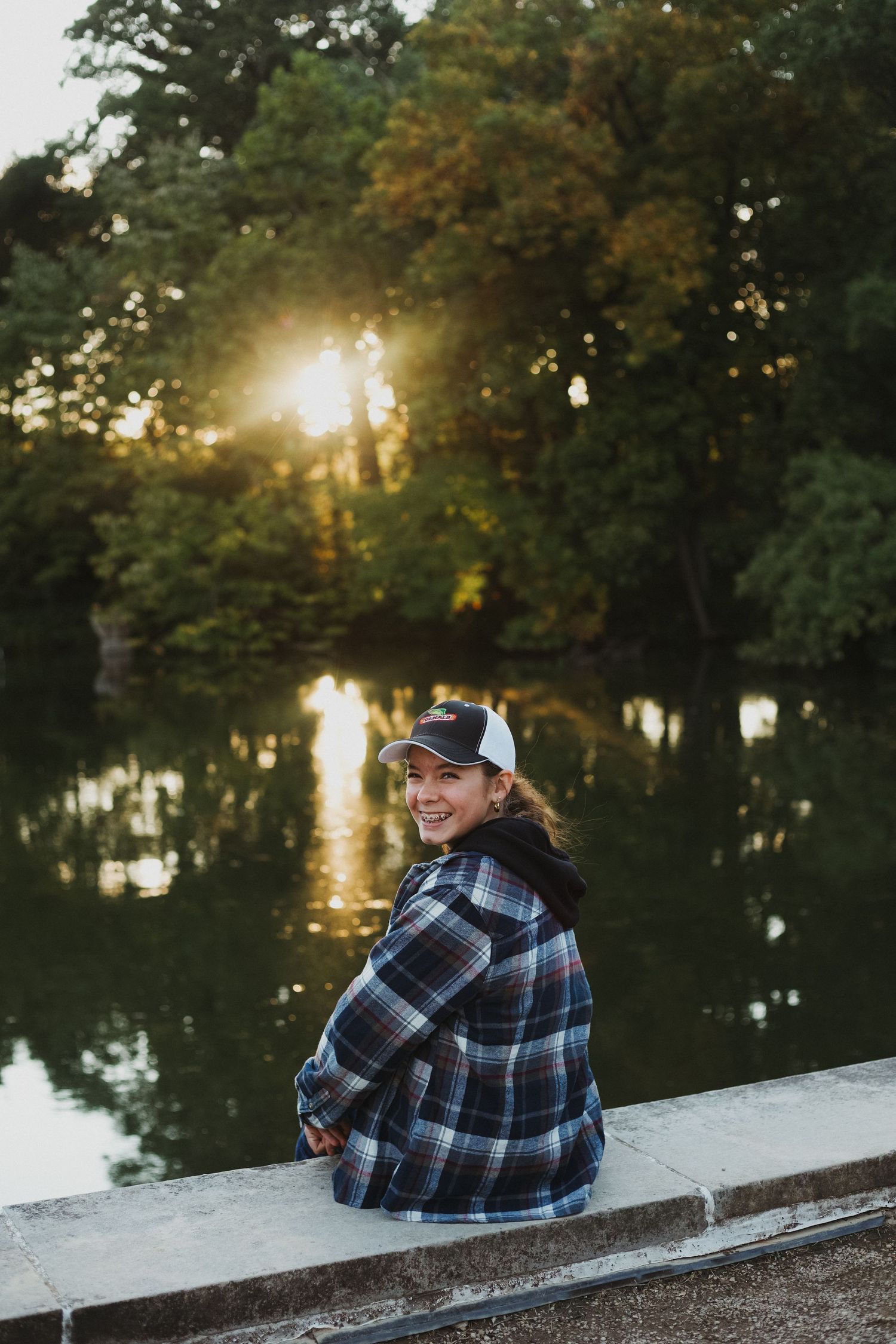Person in plaid flannel sits by pond at sunset wearing baseball cap.