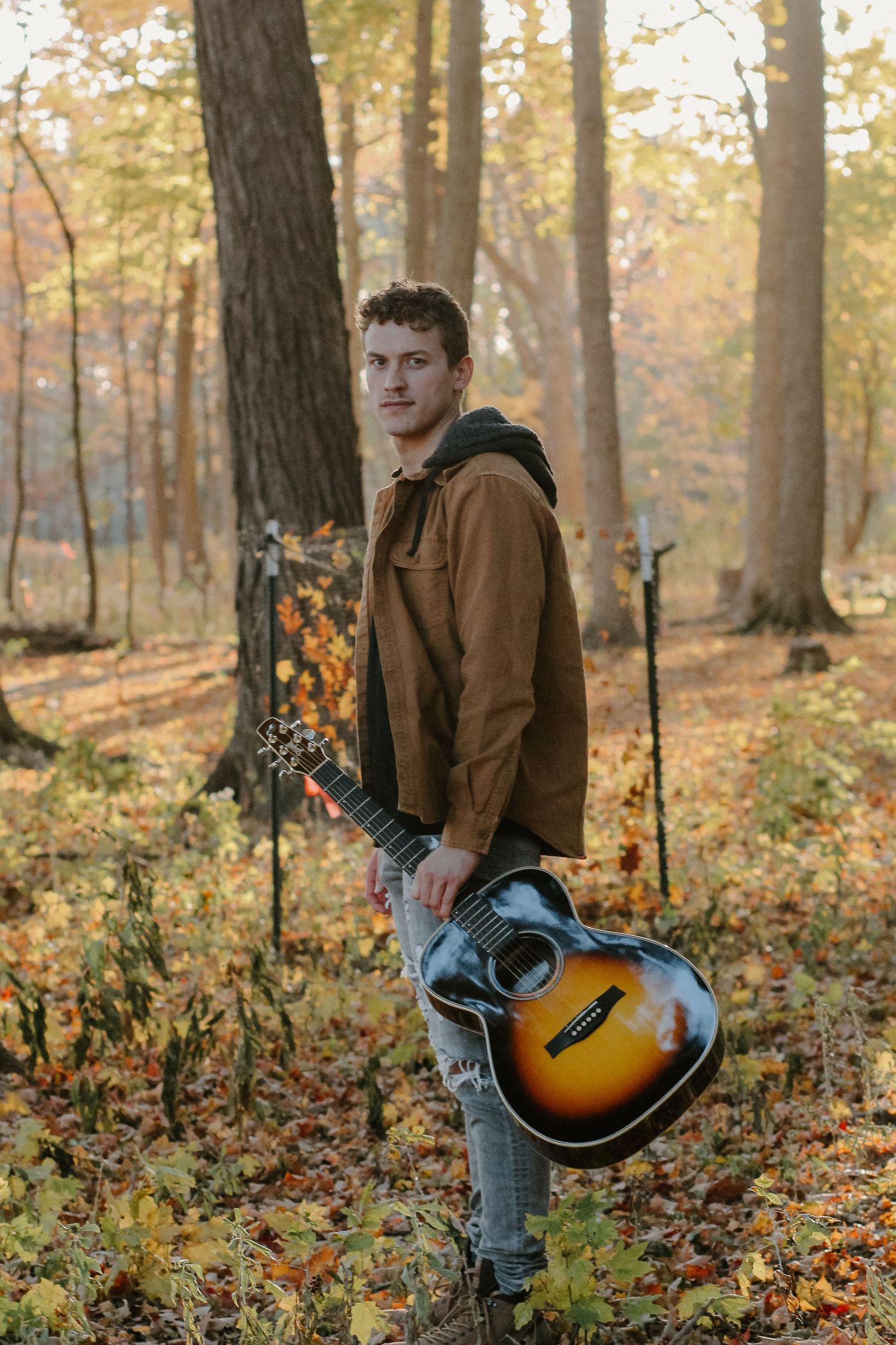 Person in brown jacket holding acoustic guitar in autumn forest setting.