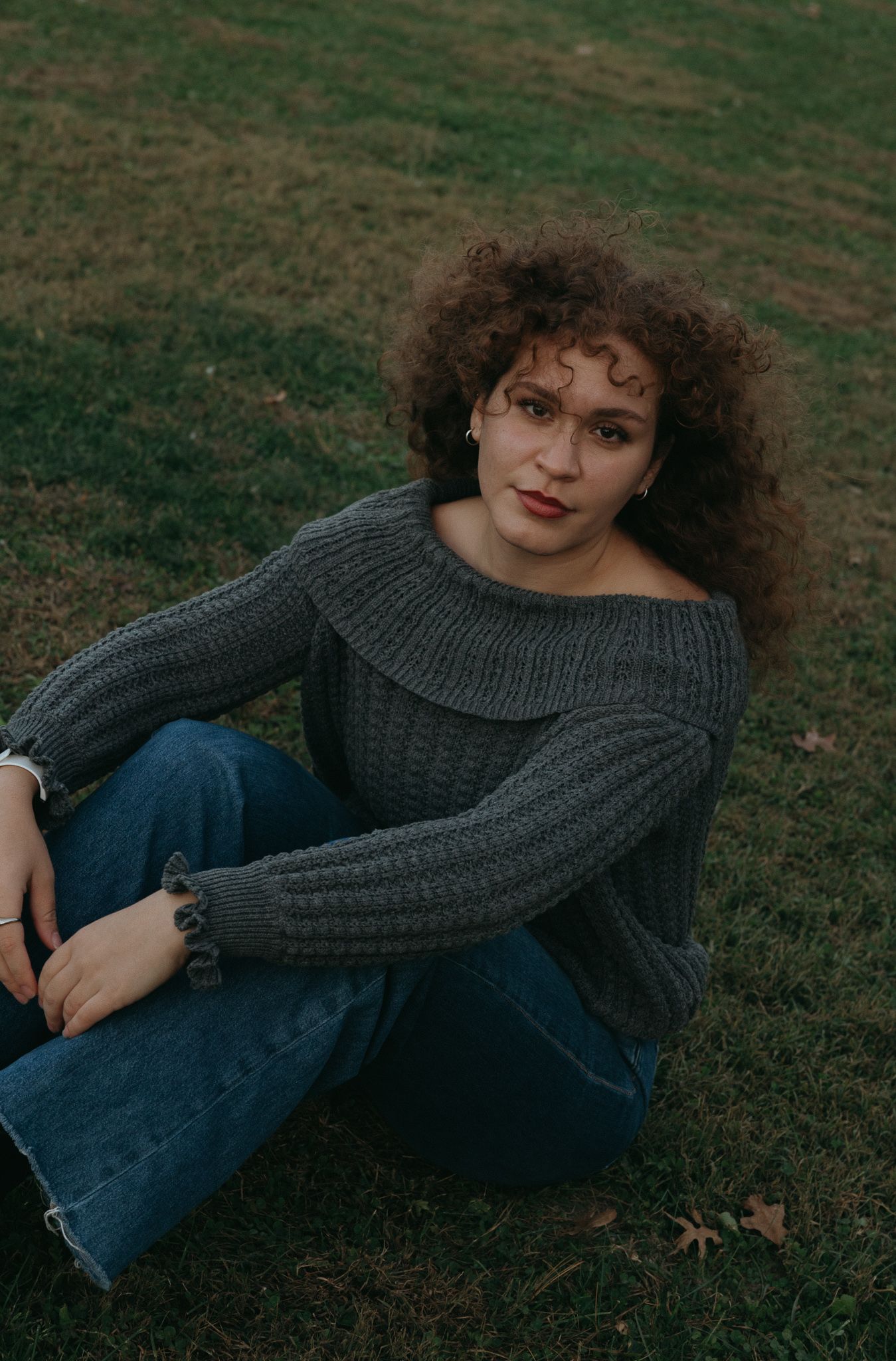 Person in gray off-shoulder sweater and jeans sitting on grass outdoors.