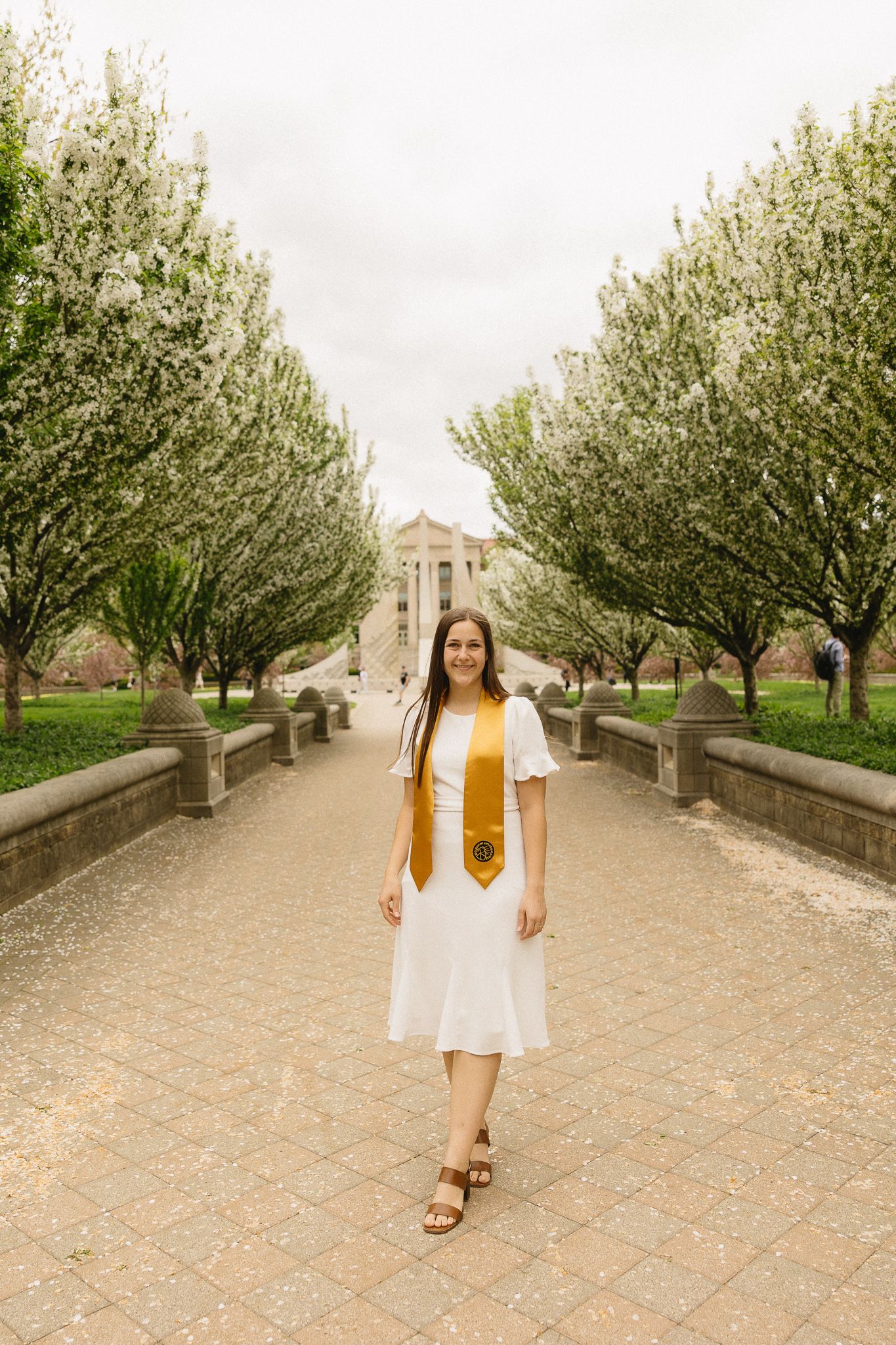 A graduate in white dress and gold stole poses on a tree-lined pathway at Purdue University.