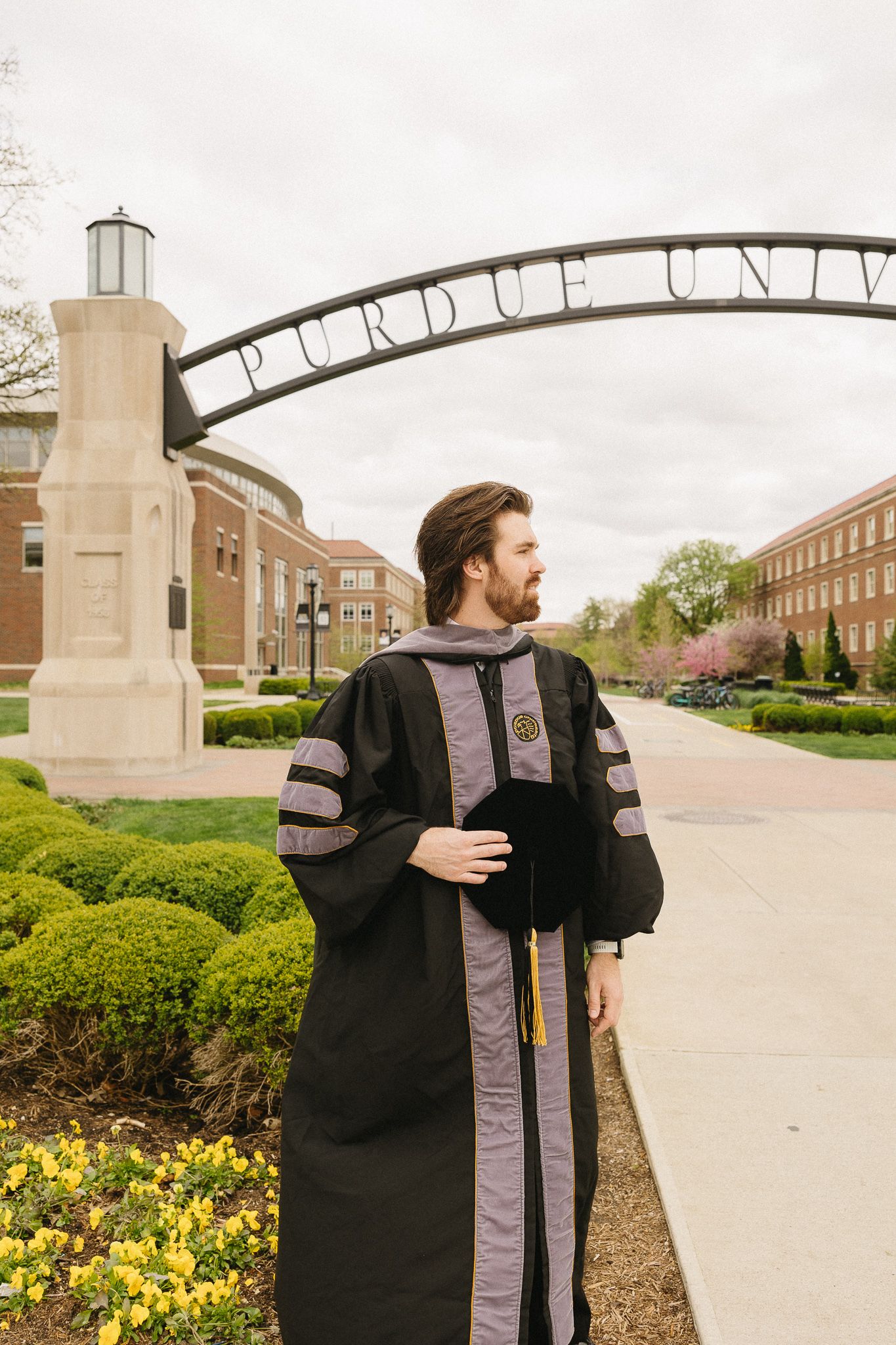 A doctoral graduate in academic regalia stands under the Purdue University entrance arch.