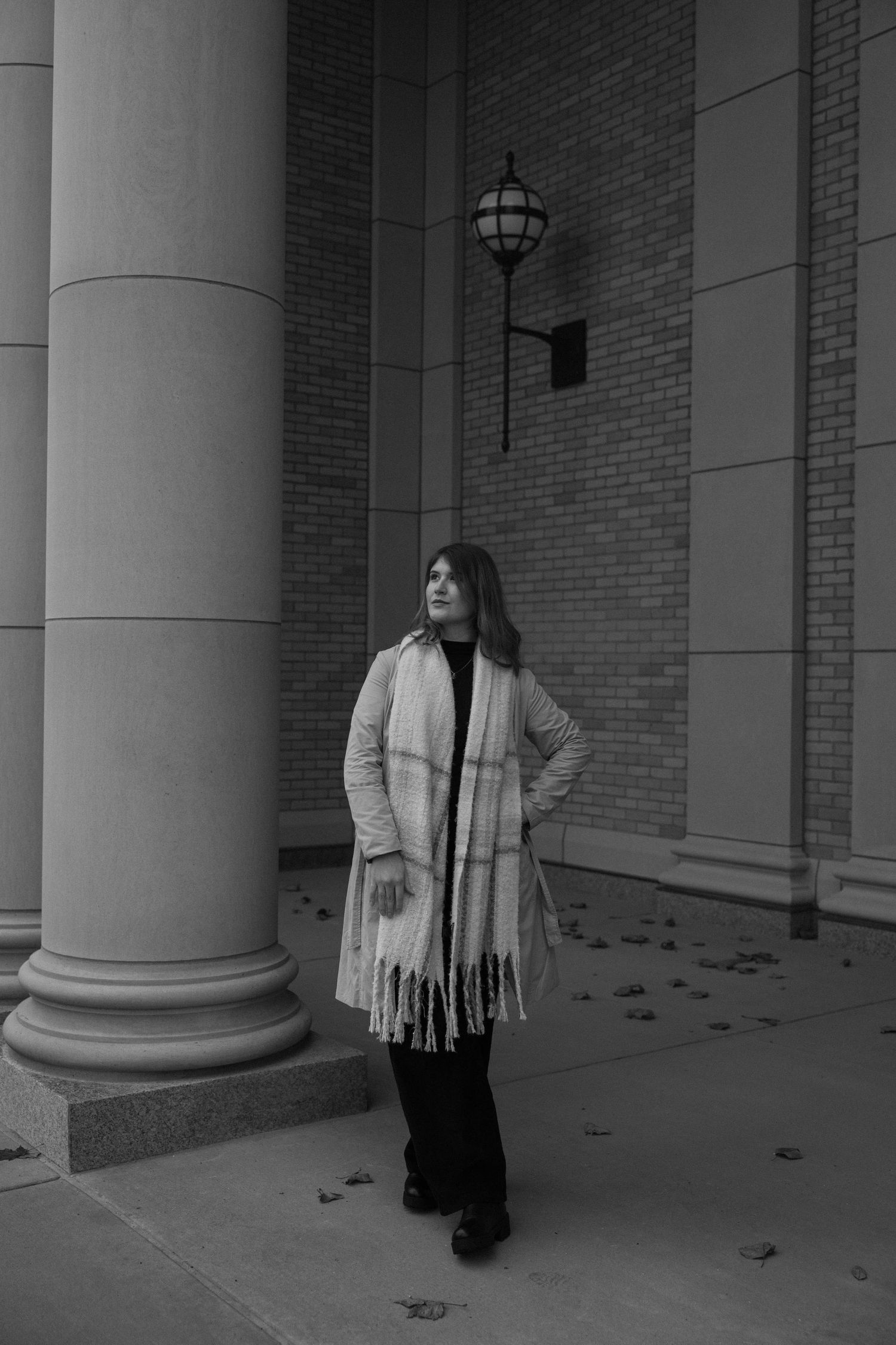 Black and white portrait of person in winter coat and scarf standing between brick columns with lantern.