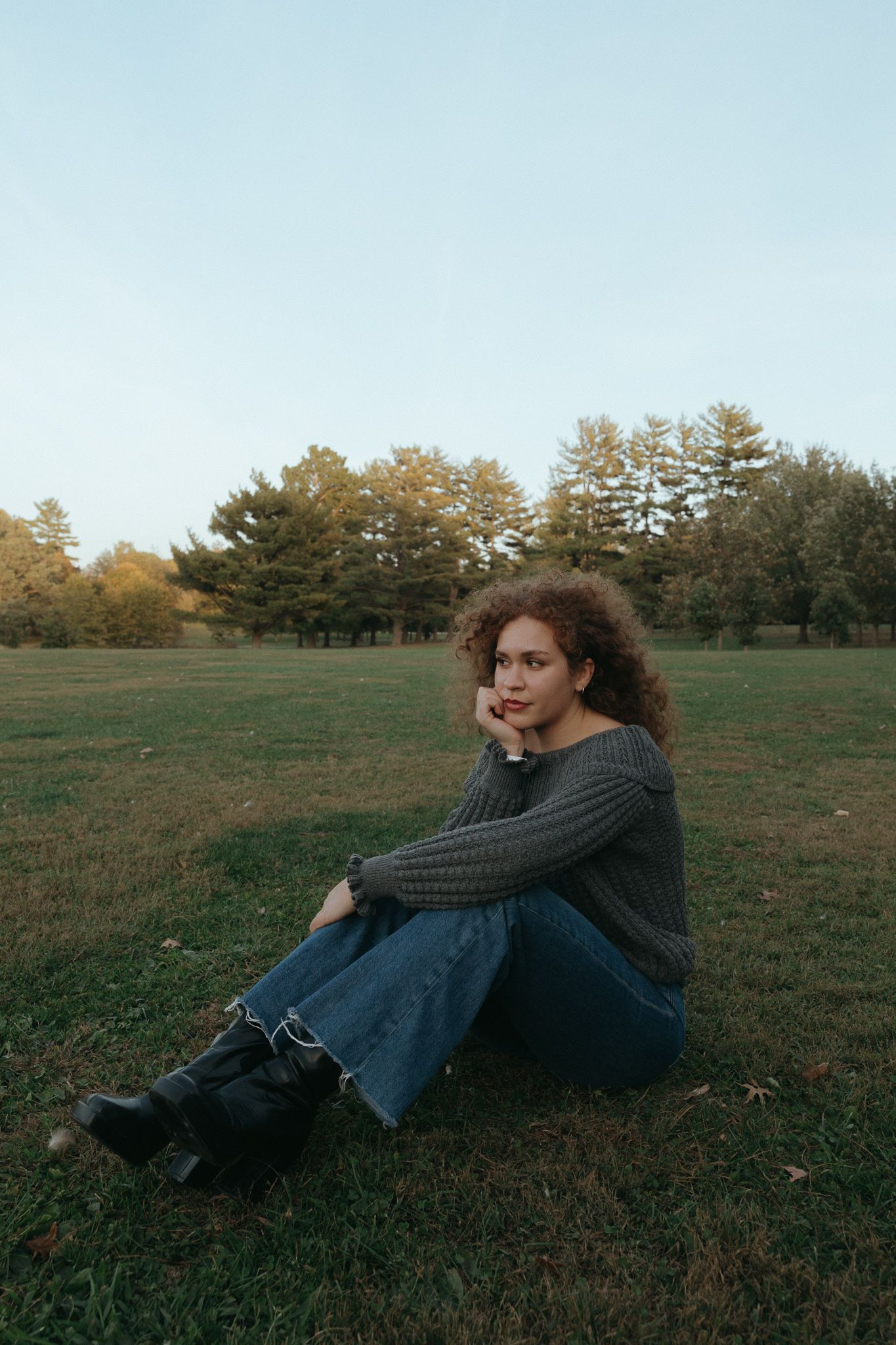 Person in cozy sweater and jeans sitting on autumn grass with trees in background.