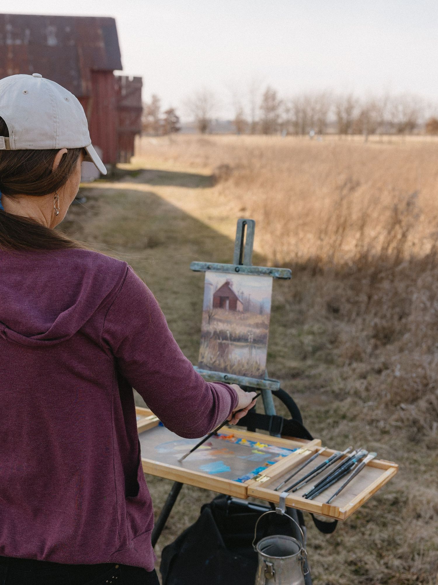 An artist paints at an outdoor easel with a rustic barn scene in the countryside.