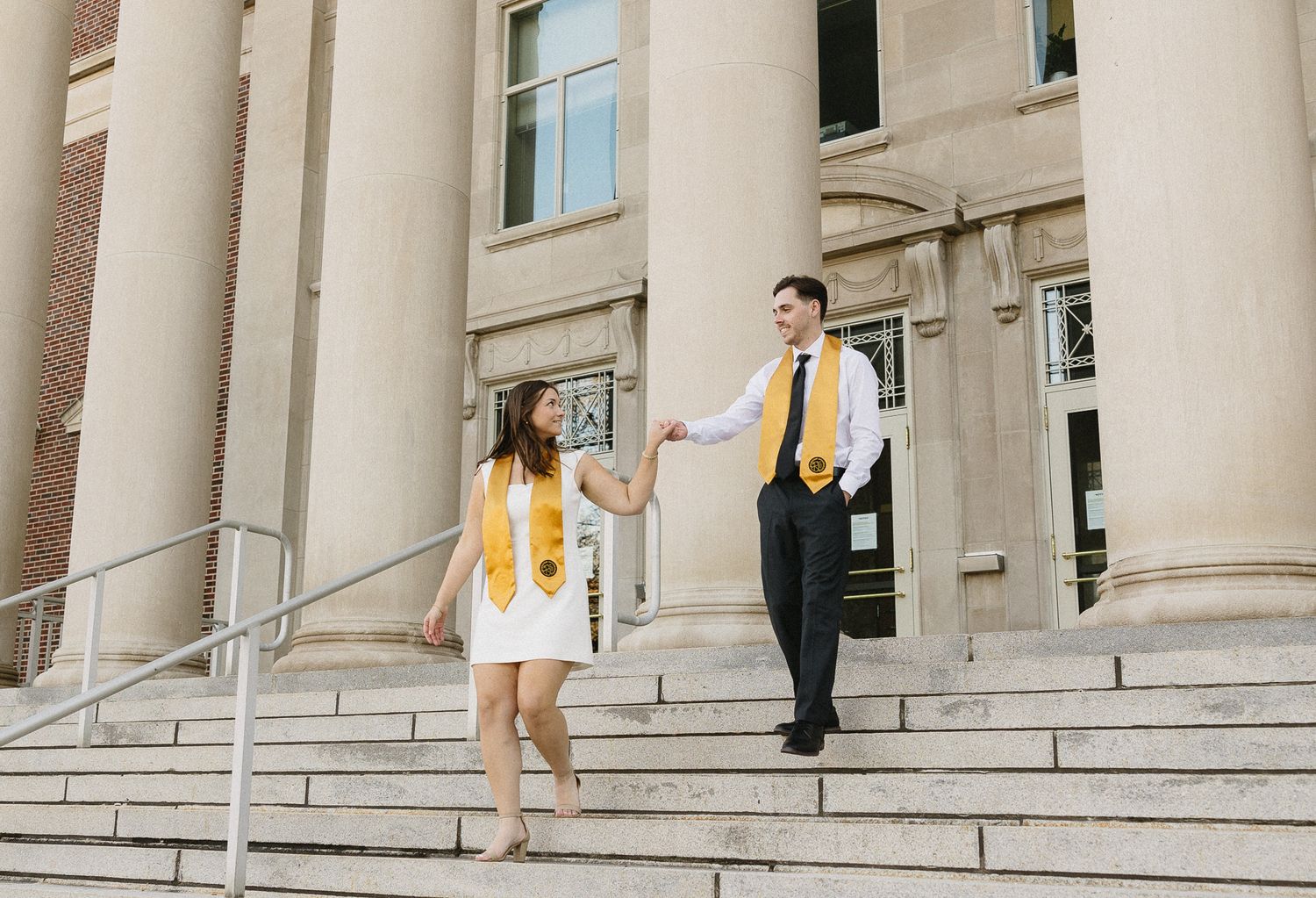 Two graduates in yellow and white attire descend stone steps of a stately building on graduation day.